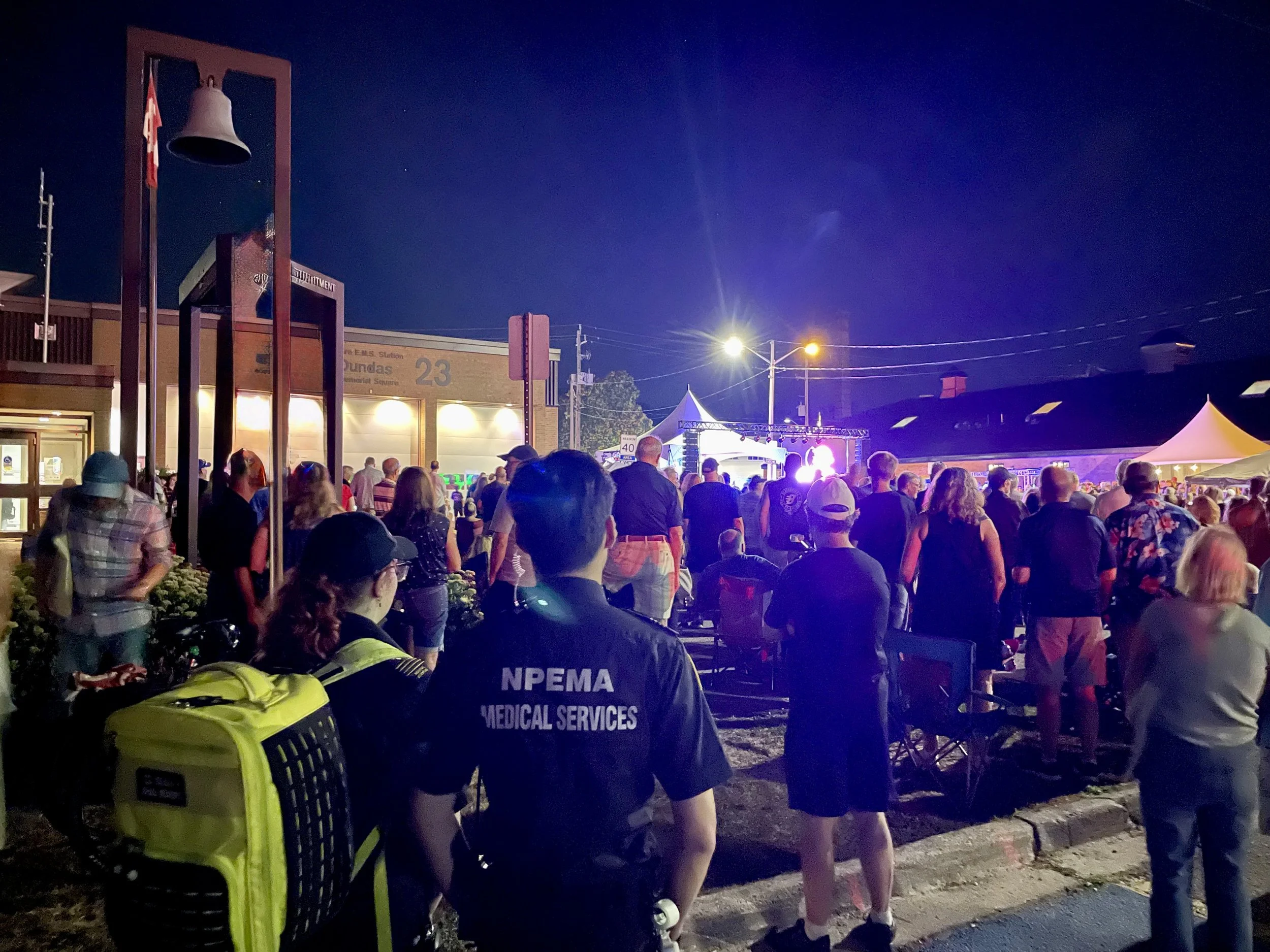 Crowd gathered at a nighttime outdoor concert beside a fire station, with stage lights, tents, and NPEMA medical services personnel.