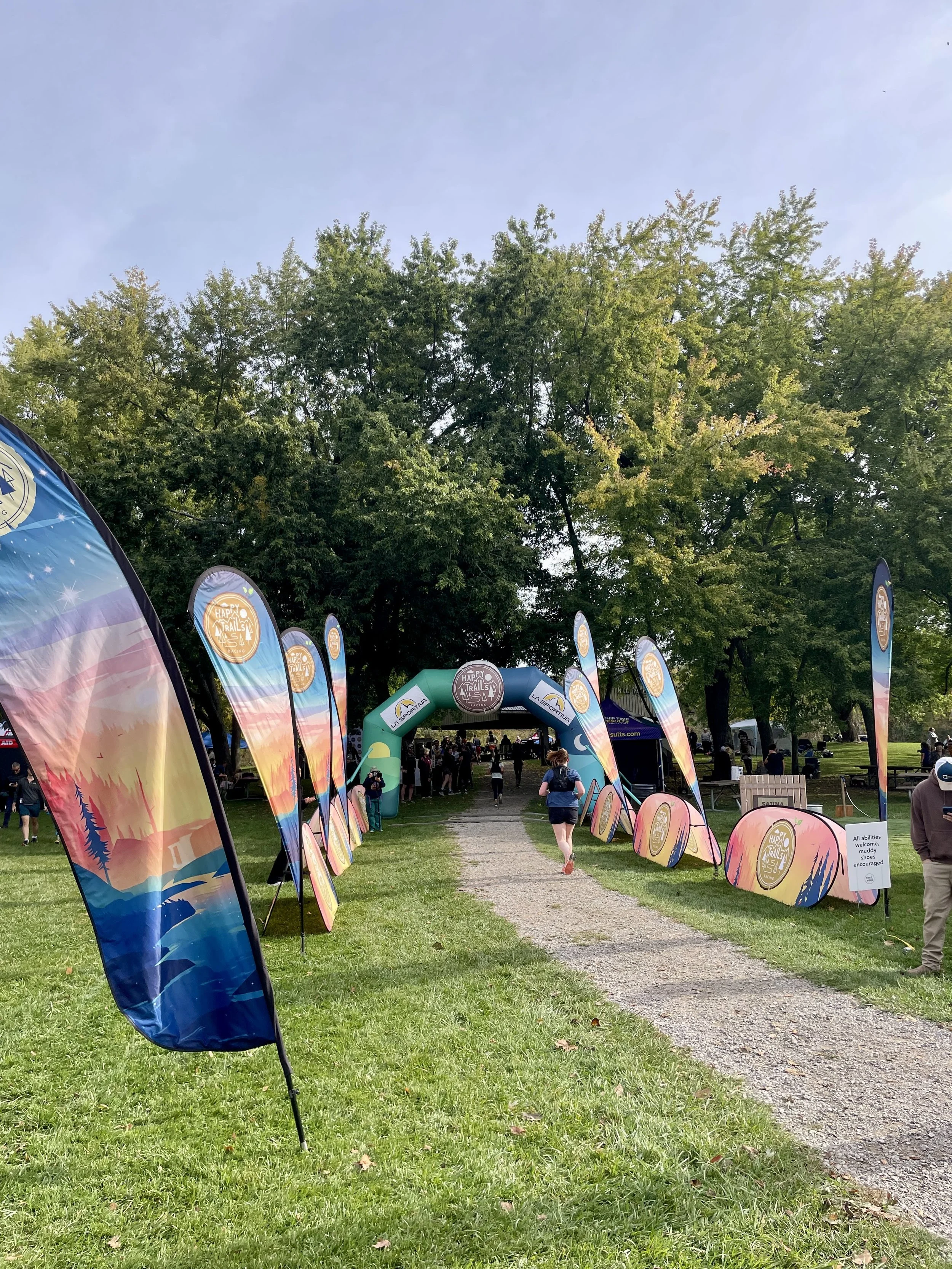 Outdoor trail race event in a park with colorful Happy Trails 5K flags, an inflatable arch, runners, and wooded surroundings.