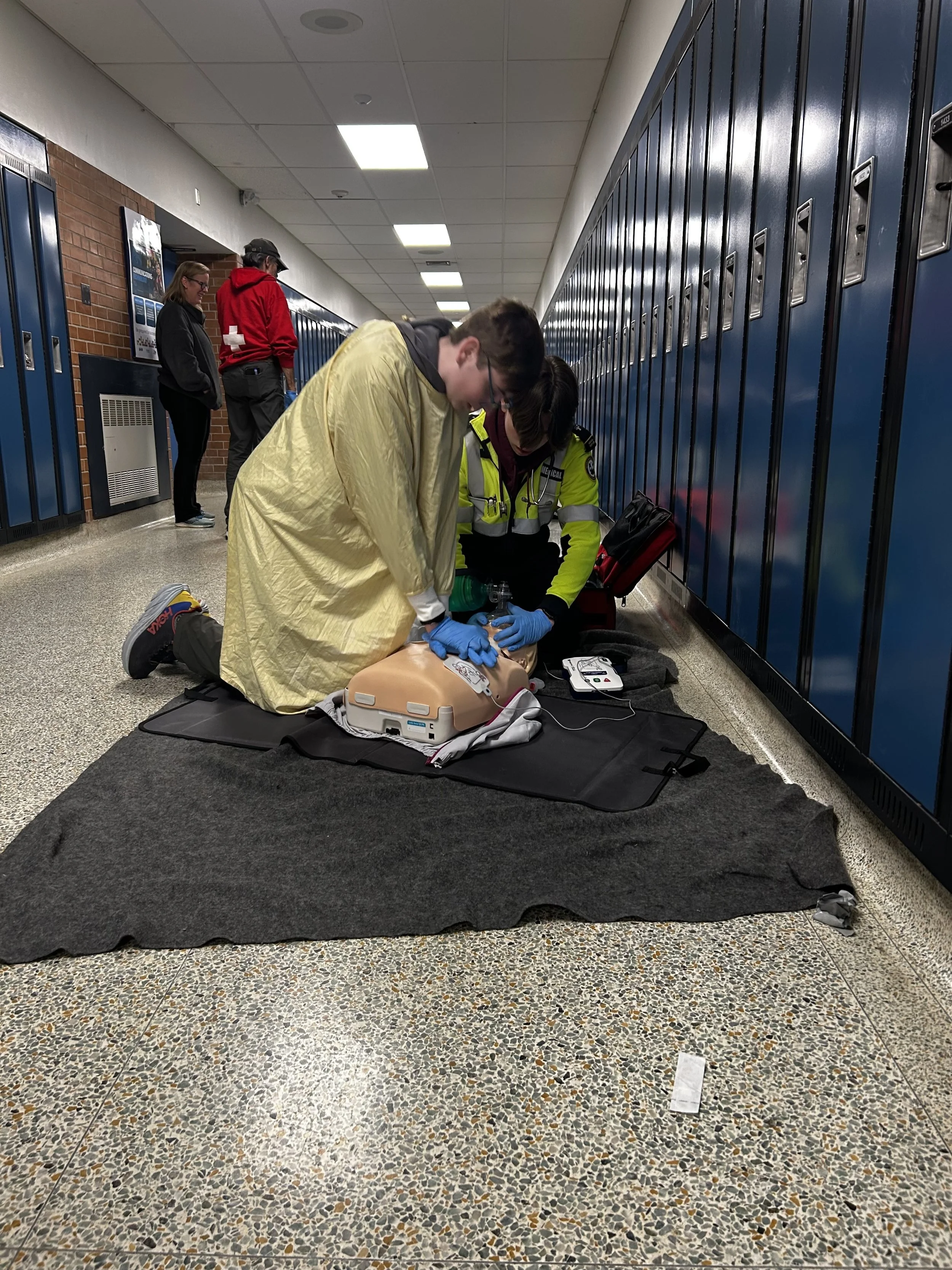 EMT performs CPR on a training mannequin in a school hallway lined with blue lockers.