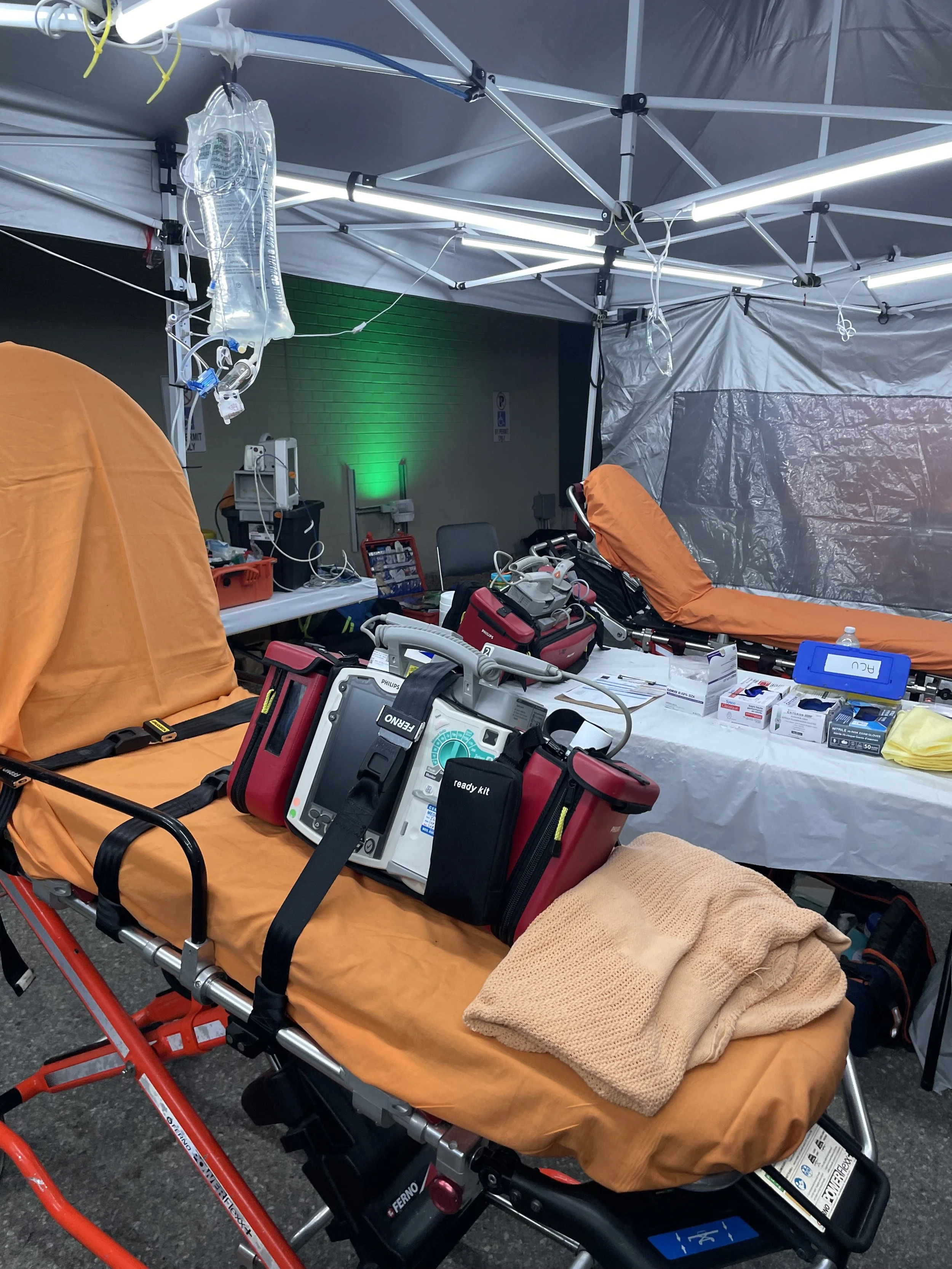 Emergency medical tent with orange stretchers, IV saline bag, defibrillator, and medical supplies.