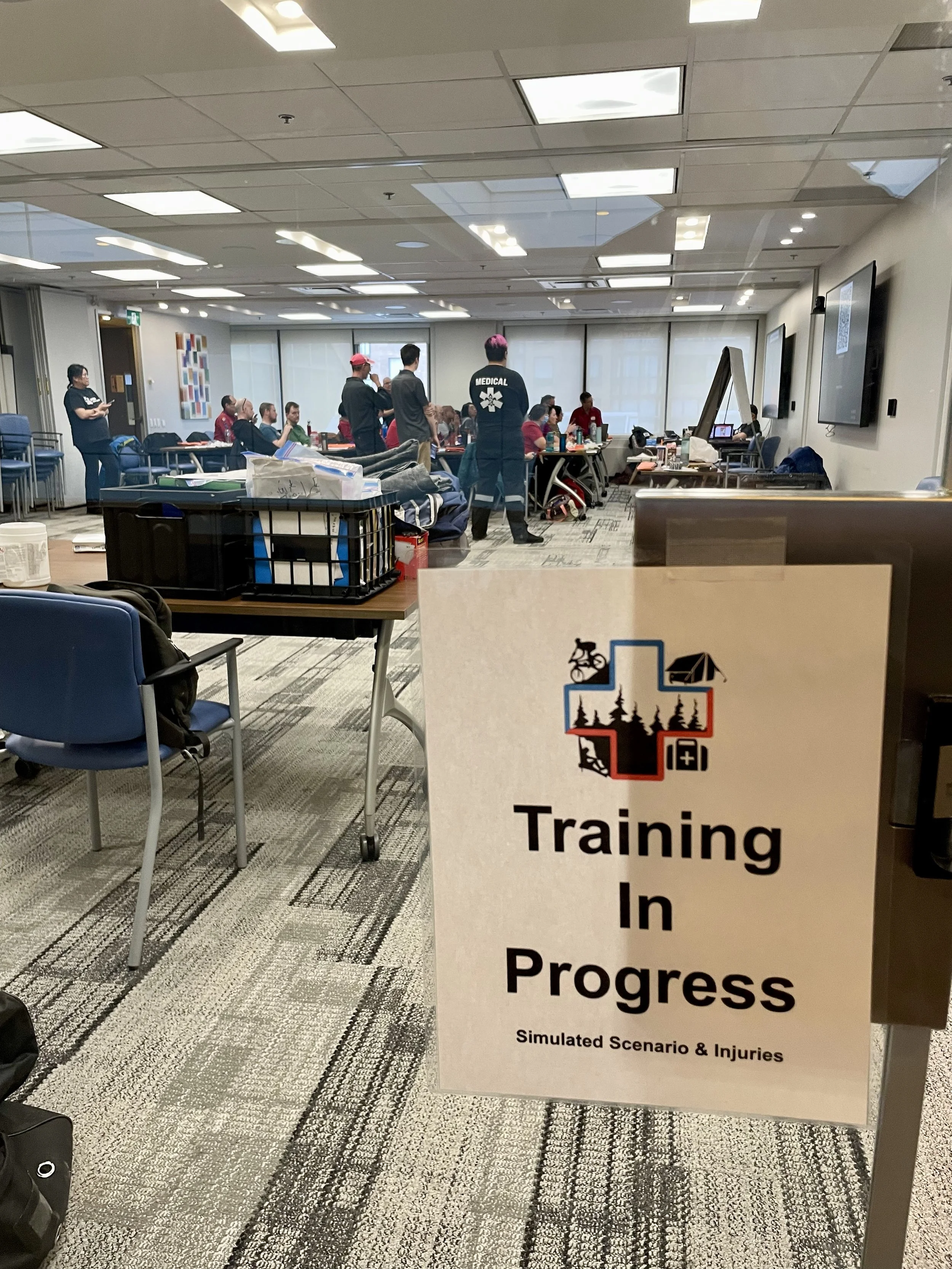 Medical training session with participants, equipment, and a sign reading “Training in Progress: Simulated Scenario & Injuries.”