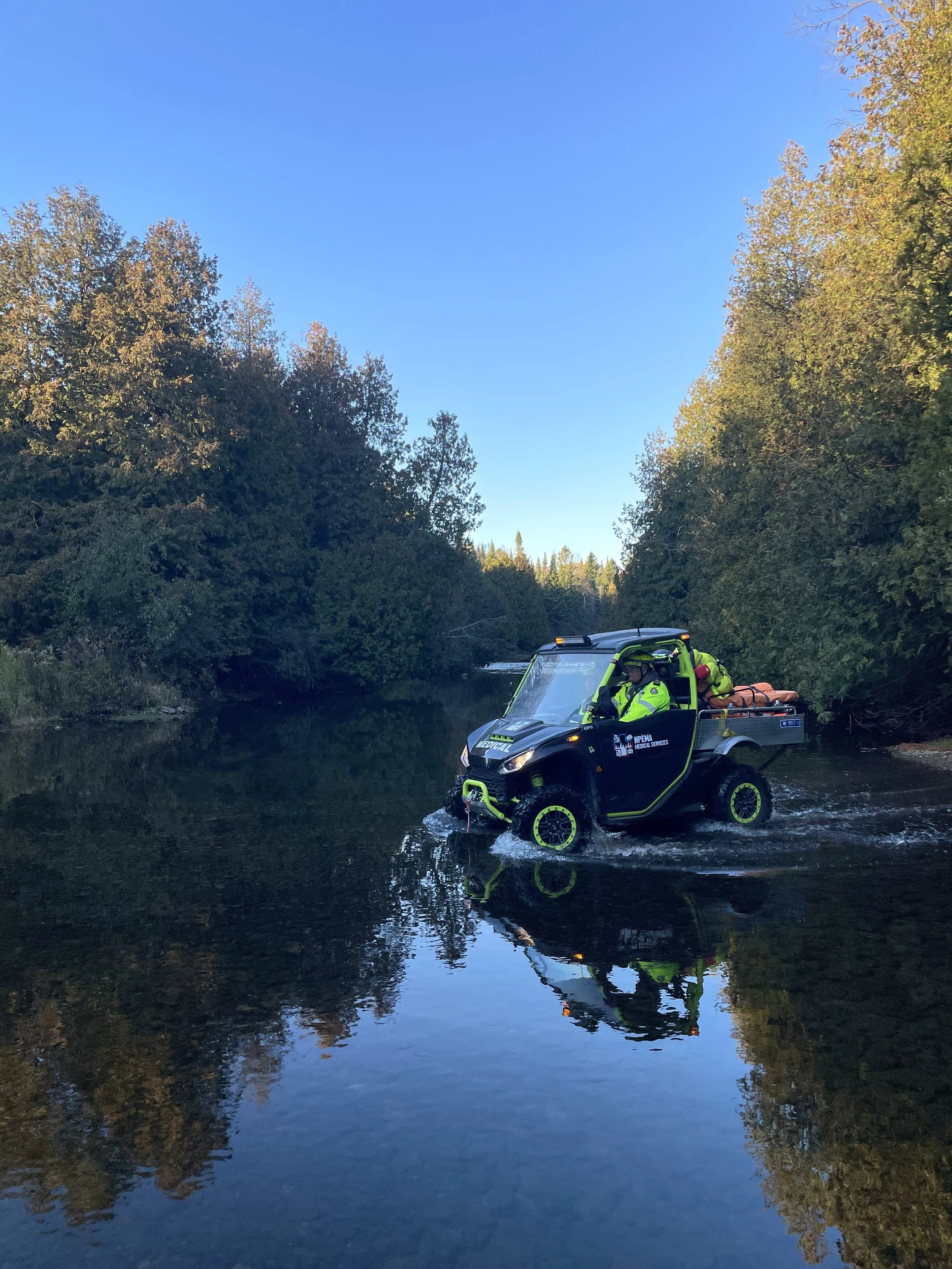 Medical rescue UTV carrying a stretcher through a shallow forest stream, surrounded by autumn trees and reflected in the water
