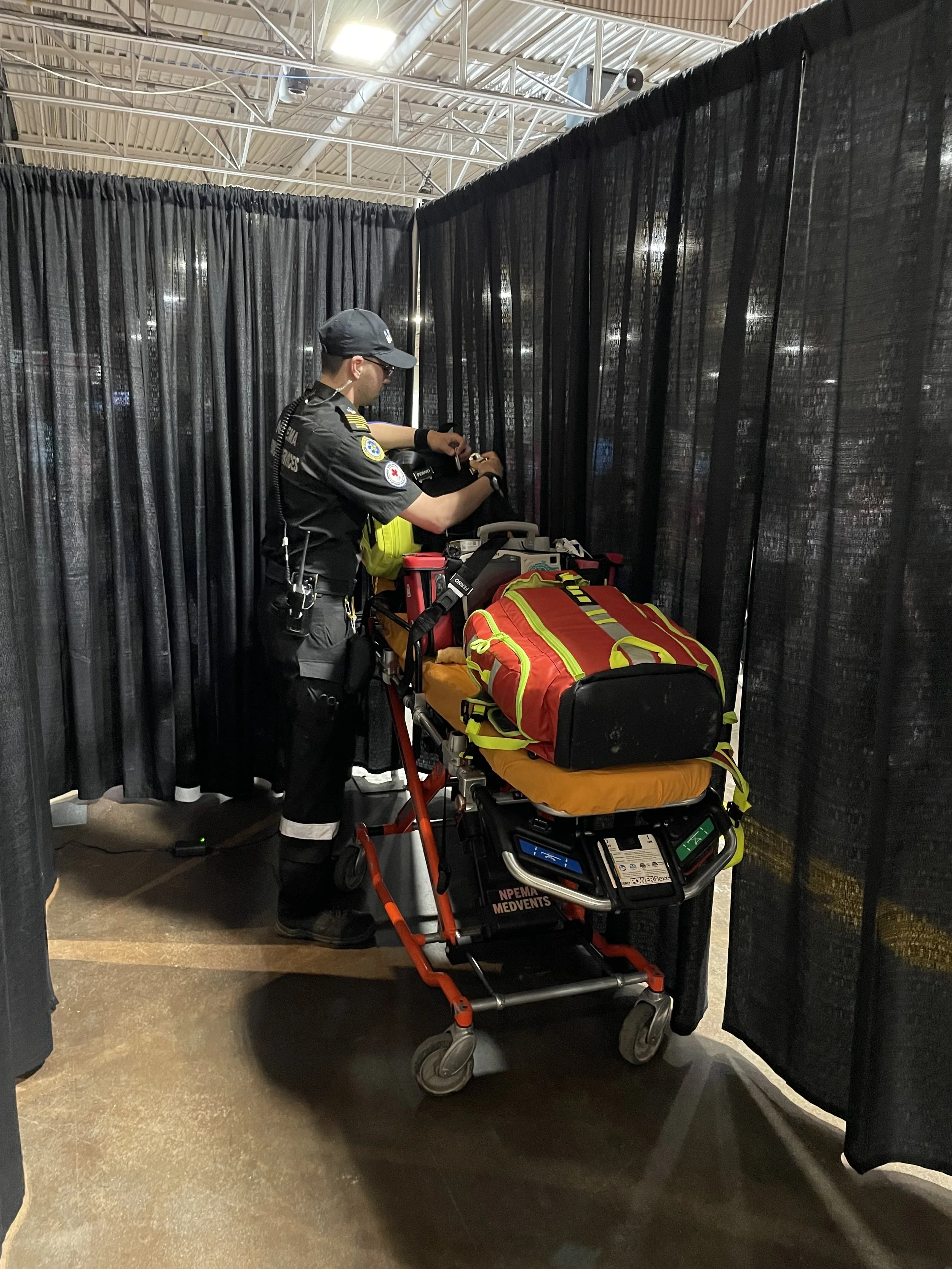 Paramedic in uniform prepares medical equipment beside a stretcher in a curtained indoor treatment area.