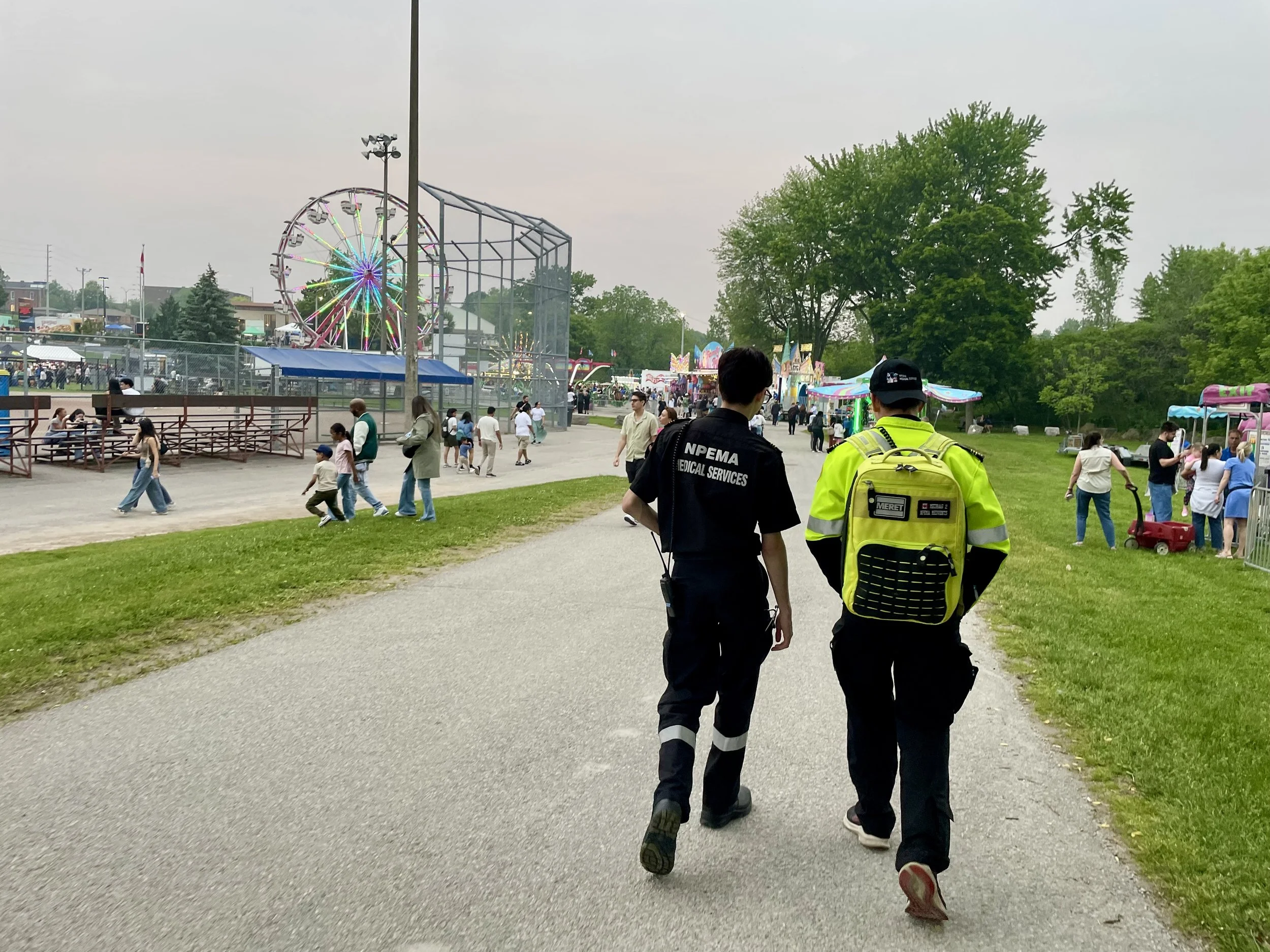 First responders walk through a busy carnival with a Ferris wheel, amusement rides, food stands, and crowds of visitors.