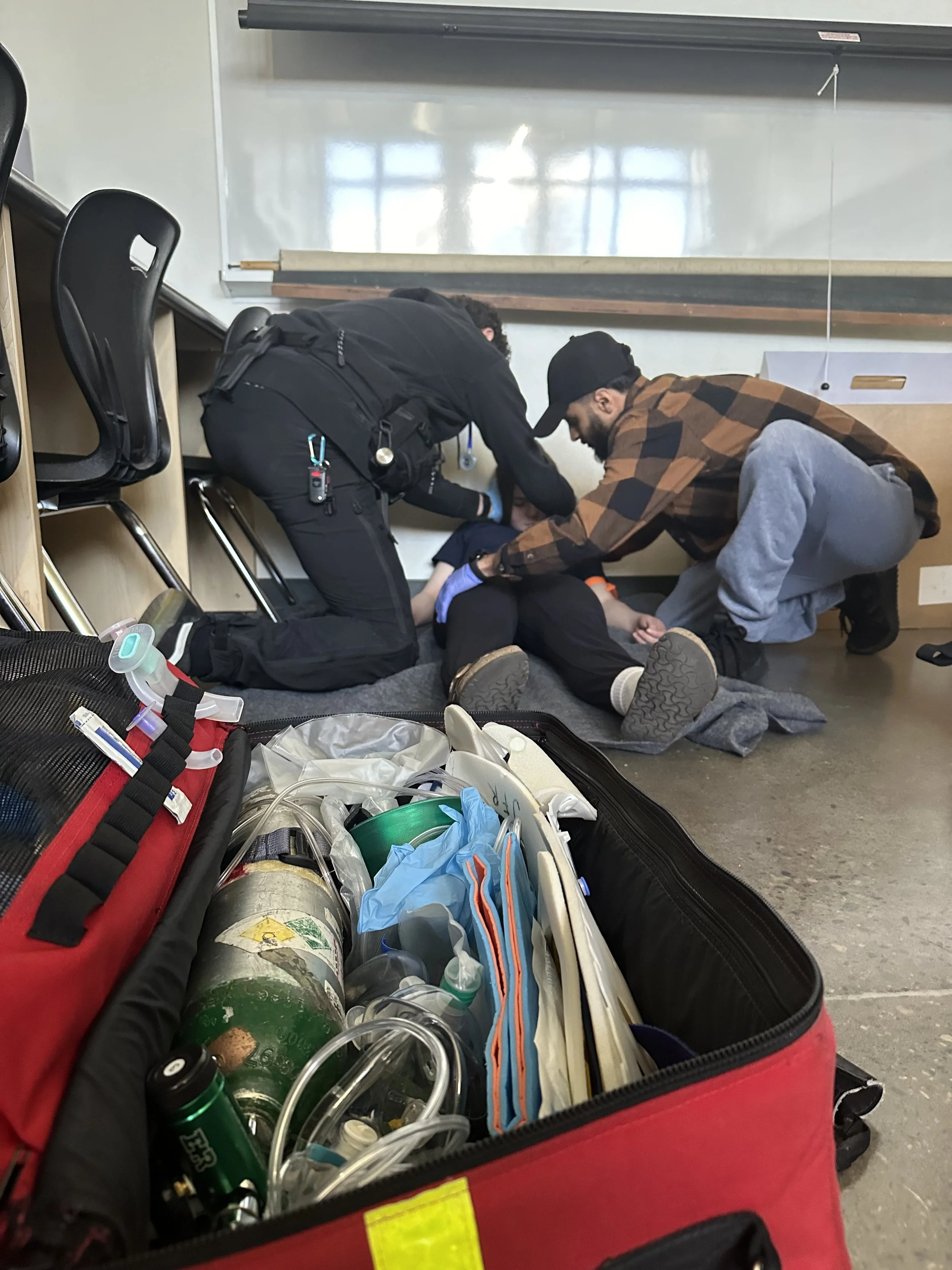 Two people provide first aid to someone on the floor beside an open medical equipment bag in a classroom.