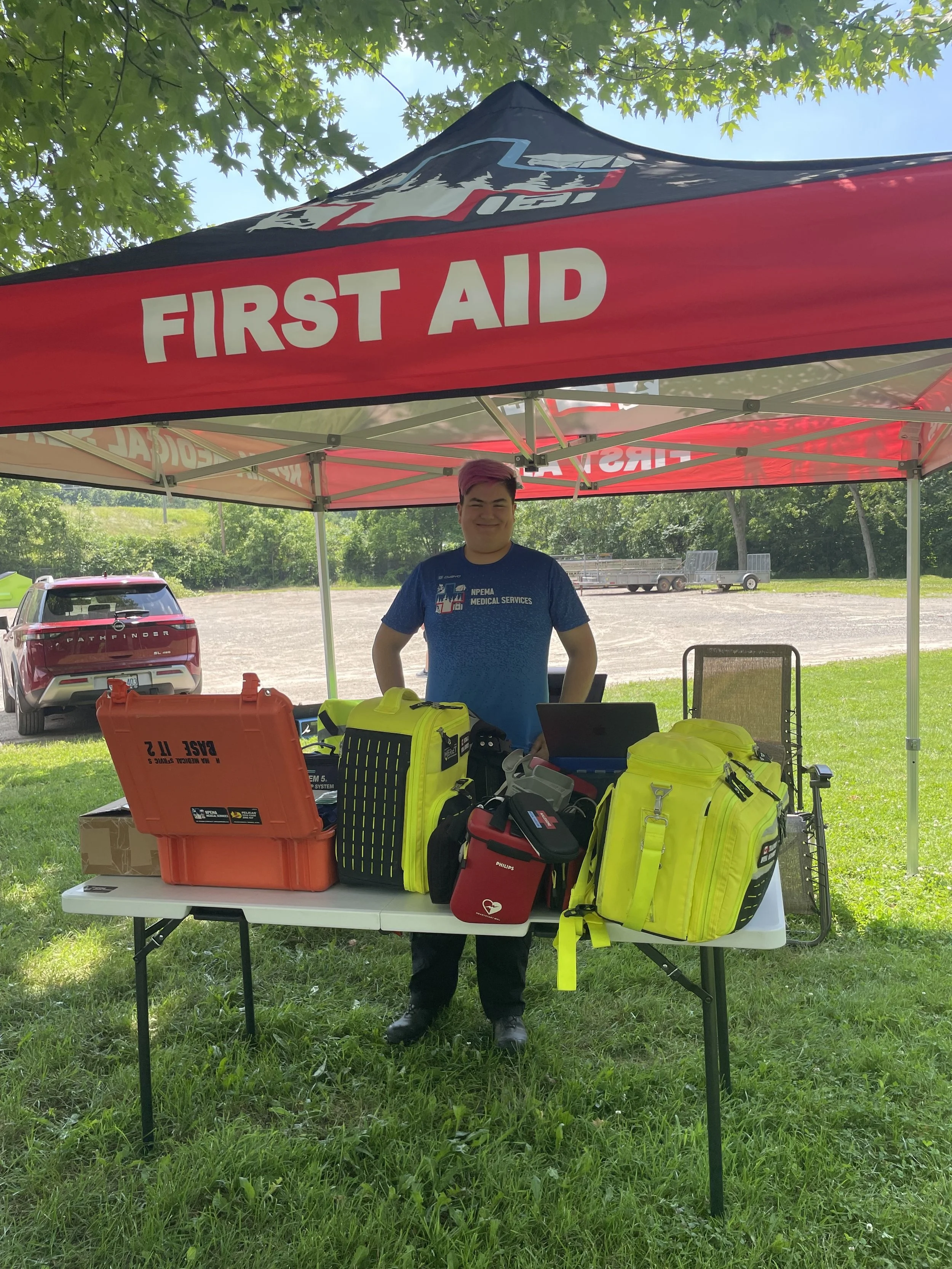 First aid volunteer standing beneath a red first-aid tent with emergency medical equipment and bags on a table.