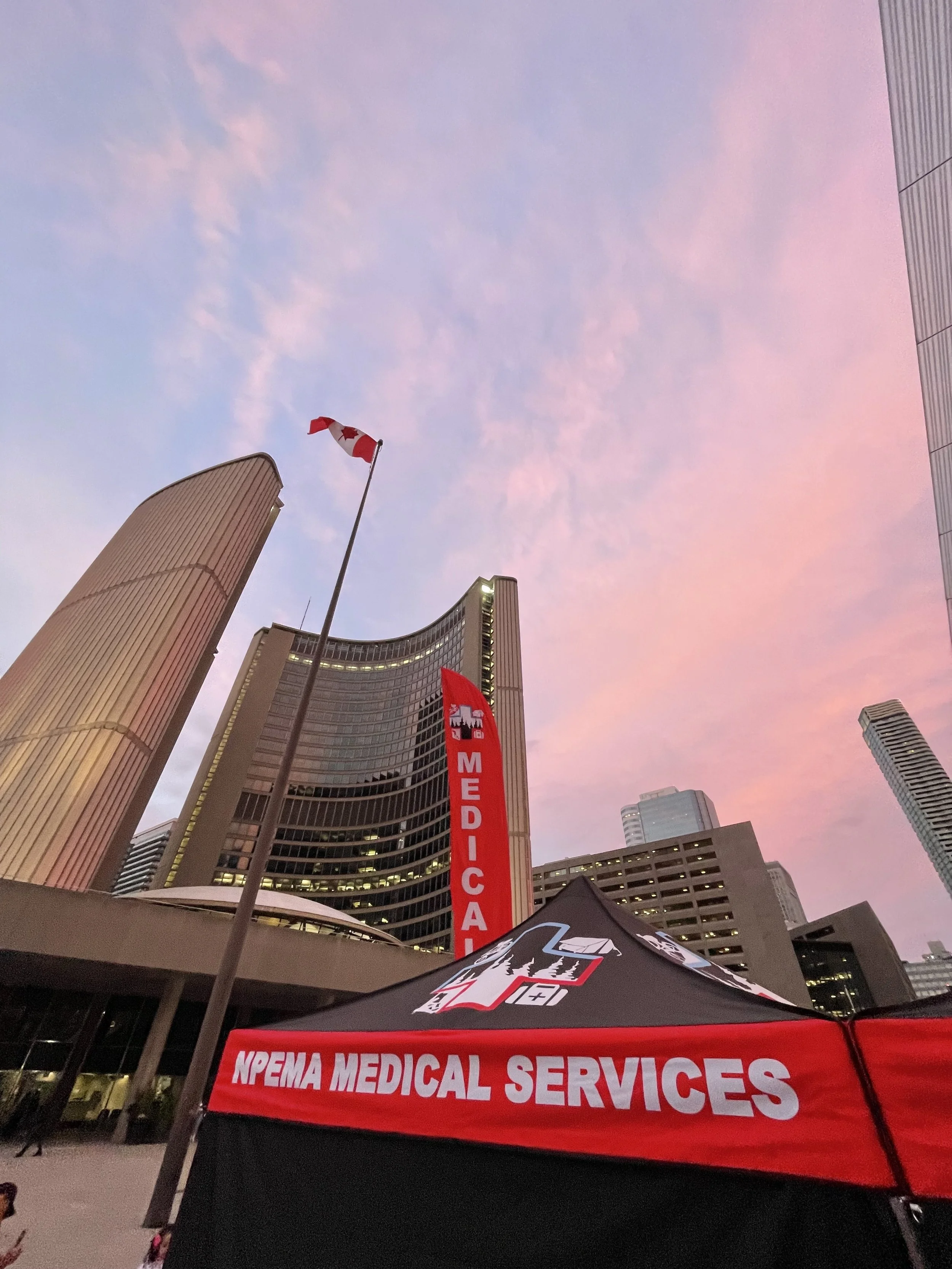 NEMA medical services tent beneath Canadian flags and Toronto City hall during sunset.