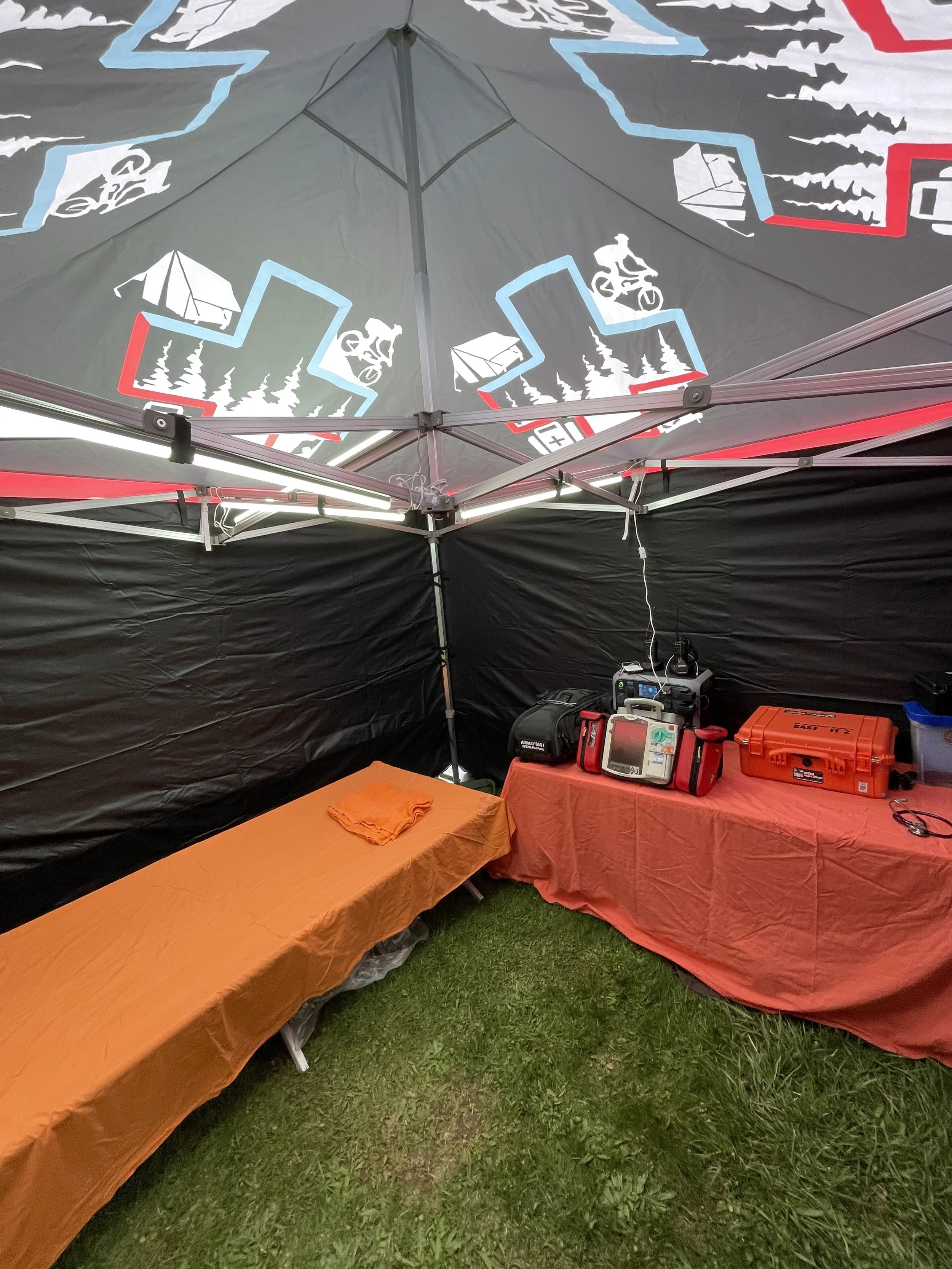 Emergency medical tent with defibrillators and medical equipment on tables, colorful patterned canopy, and grass floor