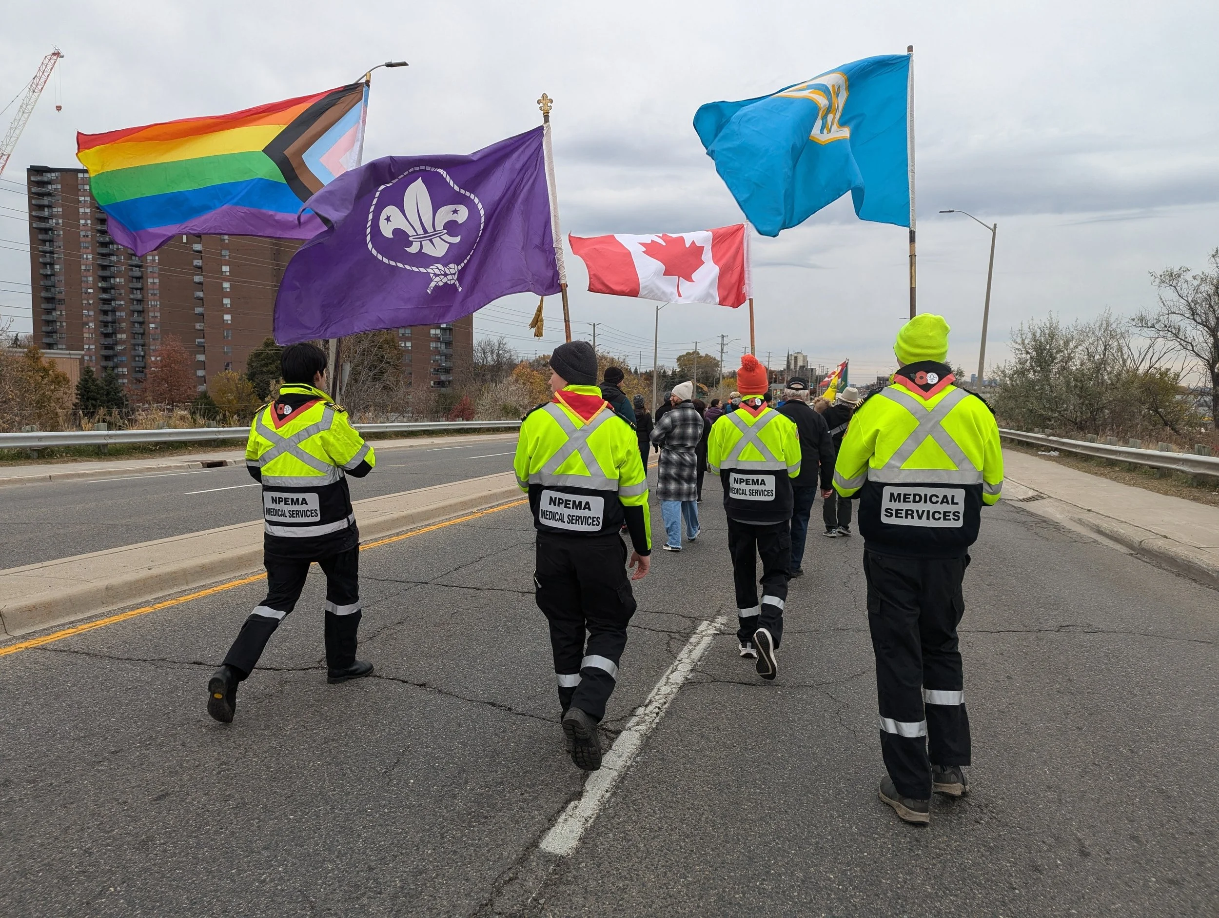 Medical service volunteers lead a street march carrying Pride, Scouts, Canada, and blue fleur-de-lis flags.