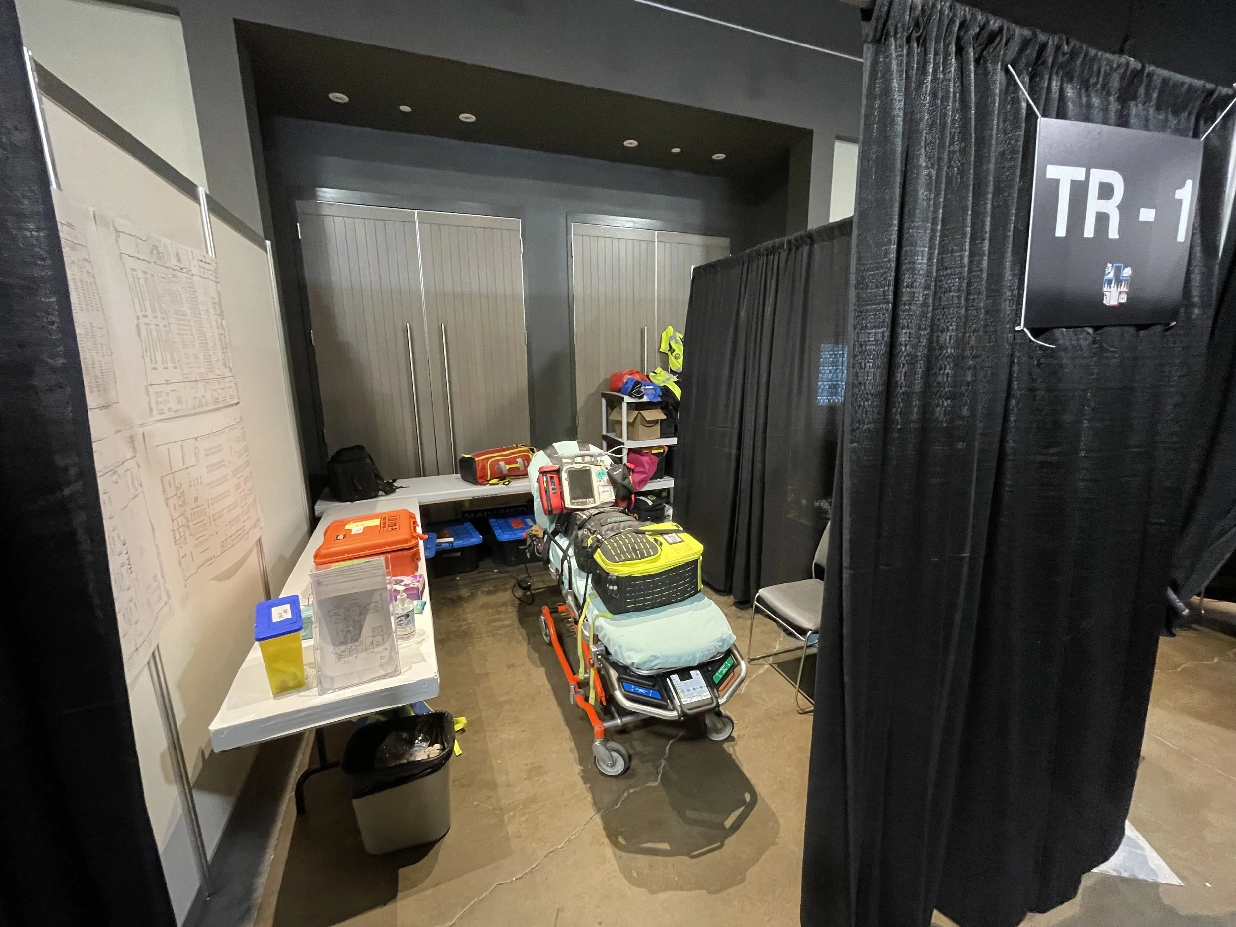 Emergency medical equipment staging area with a stretcher, medical bags, supply tables, black curtains, and a TR-1 sign