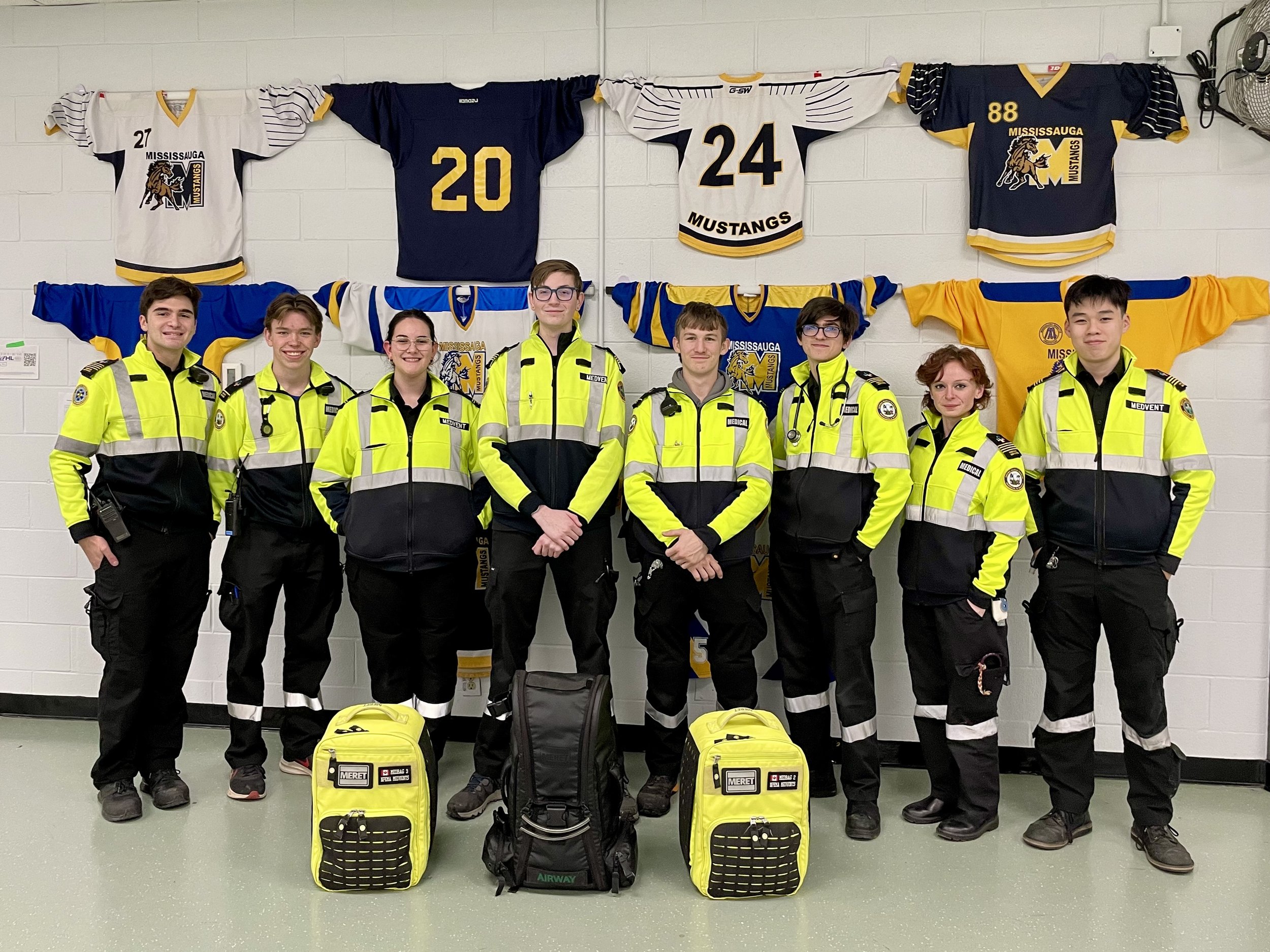 Group of eight NPEMA event medical team members in high-visibility uniforms posing with medical bags beneath Mississauga Mustangs hockey jerseys.