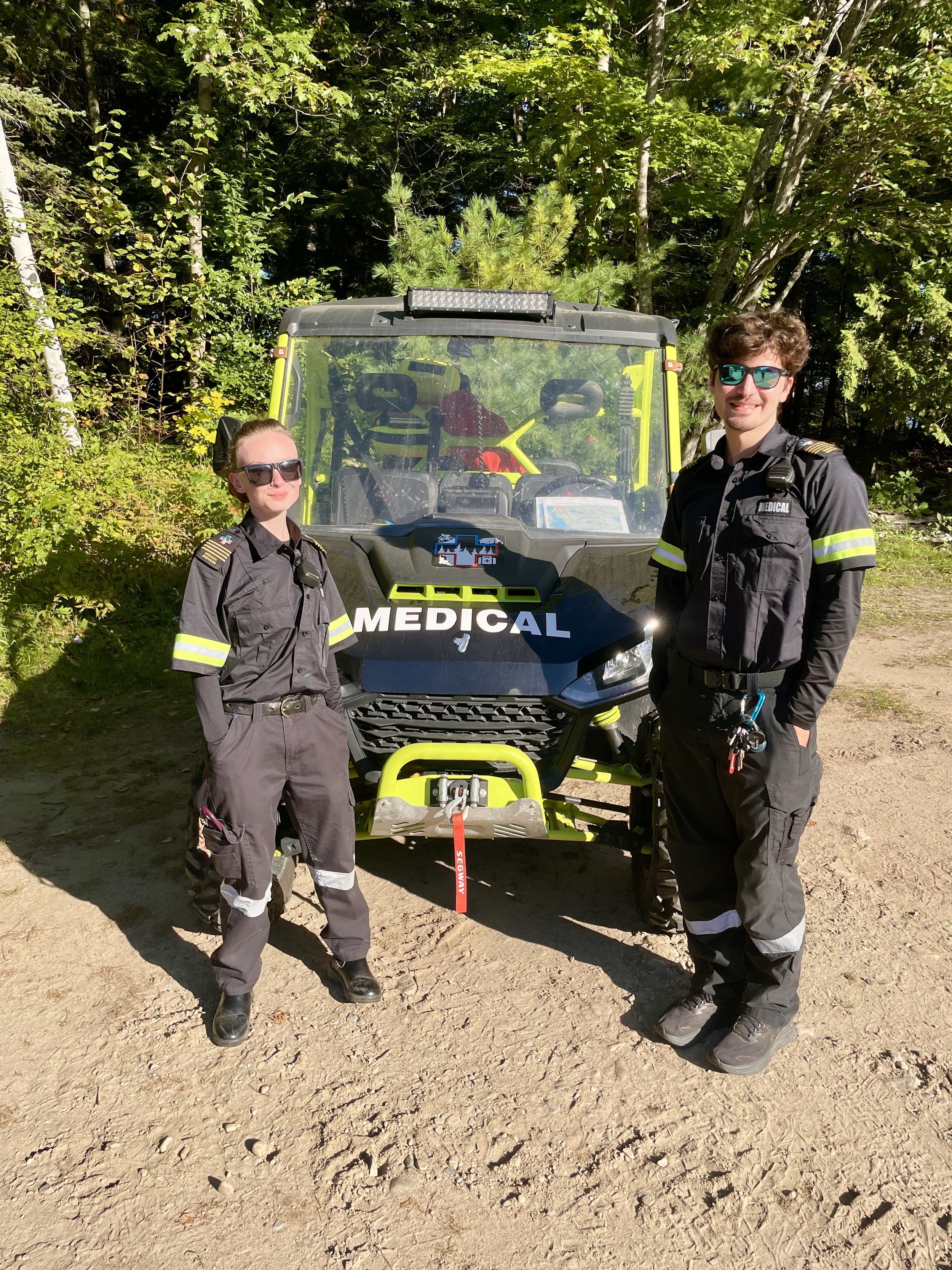 Two npema medical responders pose beside a medical off-road utility vehicle on a forest trail.