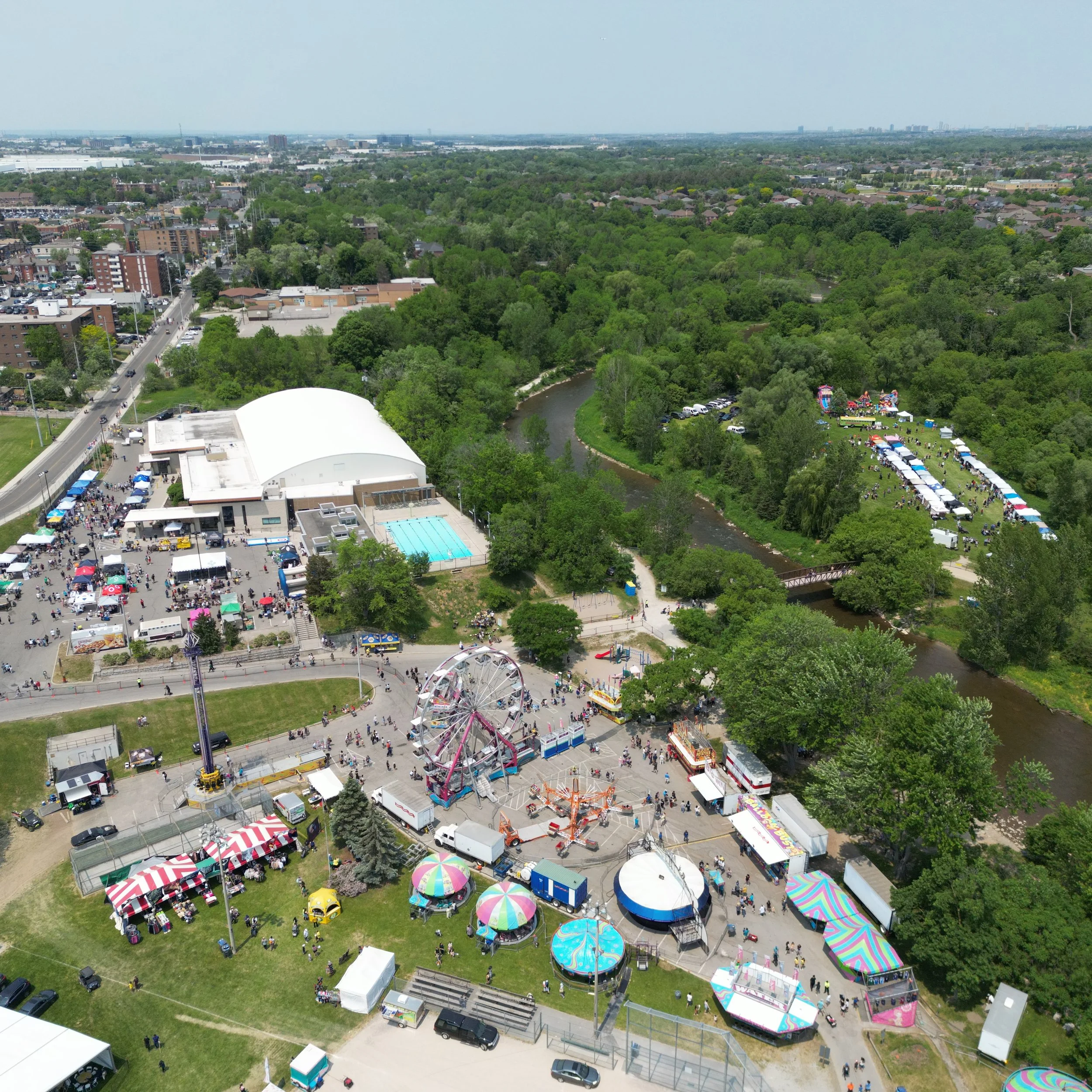 Aerial view of a bread & honey city park festival with carnival rides, food tents, a swimming pool, and a river surrounded by trees and neighborhoods.