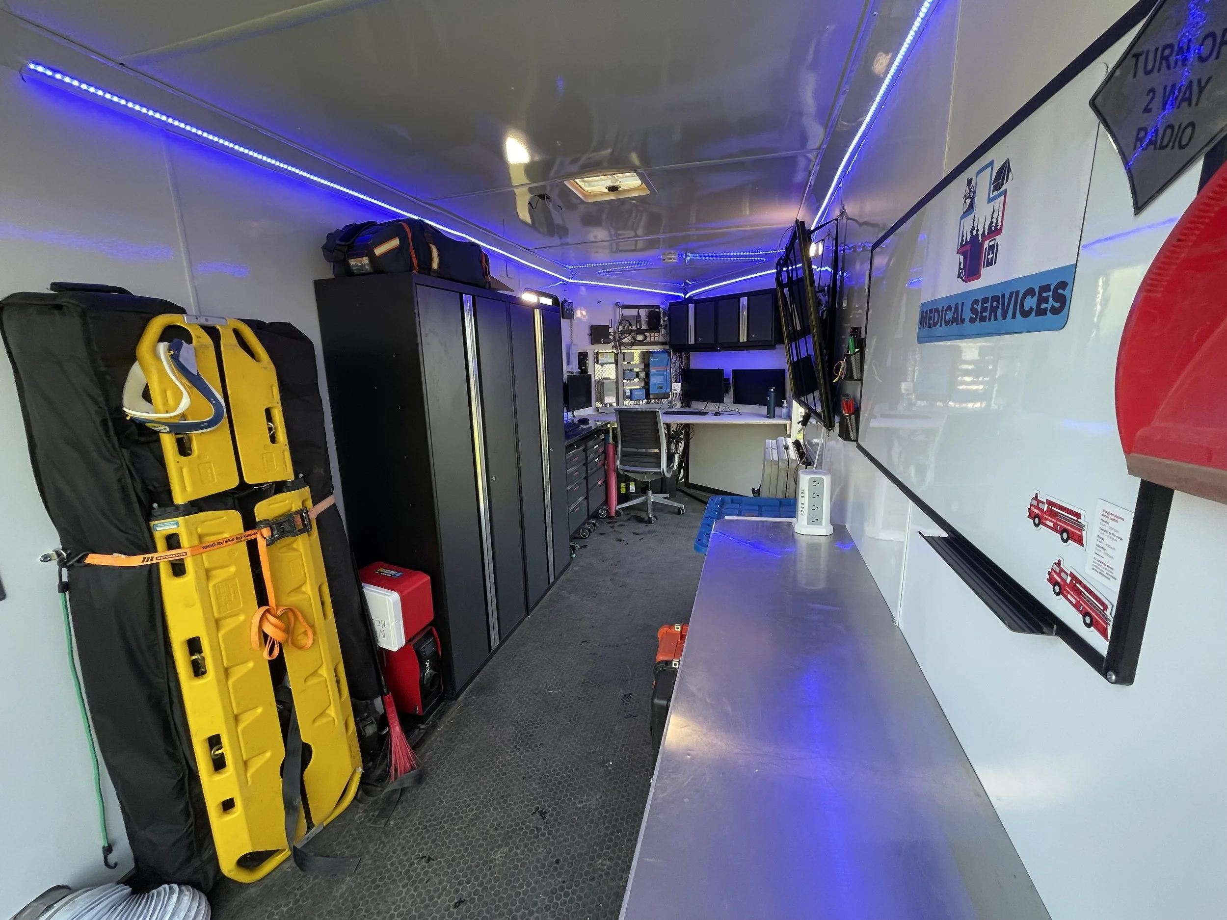 Interior of a mobile medical services ambulance with a yellow spine board, emergency equipment, cabinets, workstations, and blue LED lighting