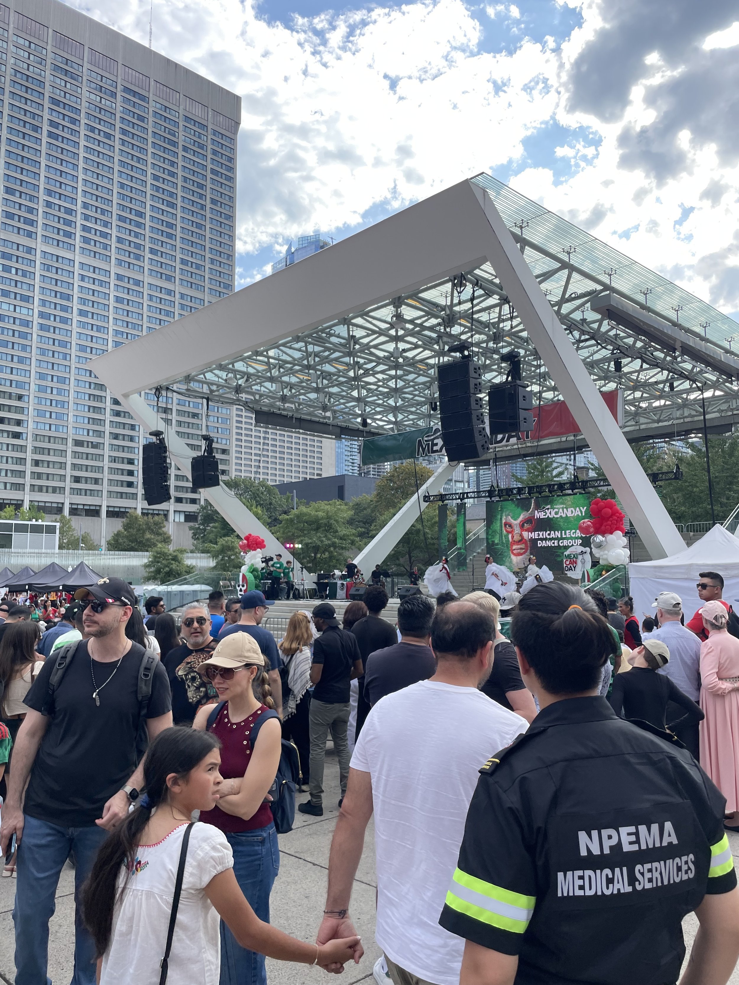 NPEMA medical responder and crowd watching a Mexican Day cultural festival with dancers on an outdoor stage beneath a glass canopy in downtown Toronto