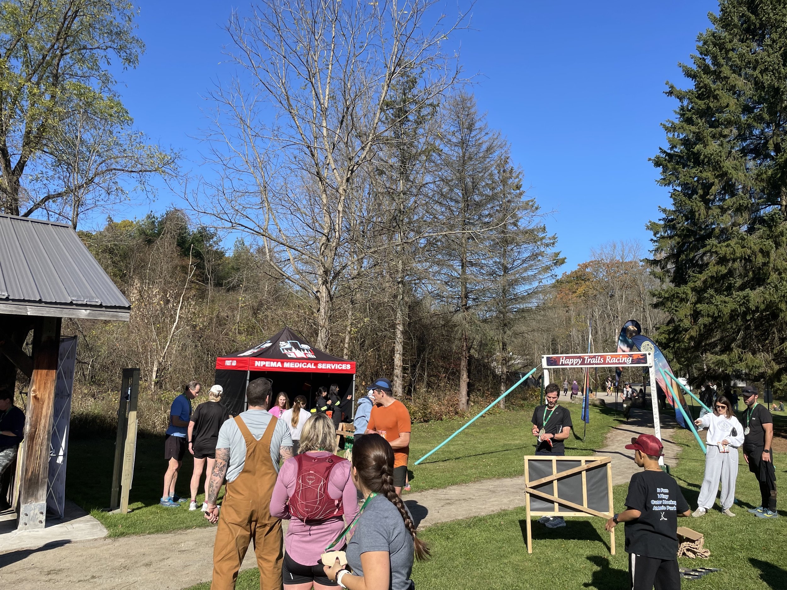 Runners gather at an outdoor trail race event beneath a Happy Trails Racing arch, with a medical services tent and wooded park surroundings.