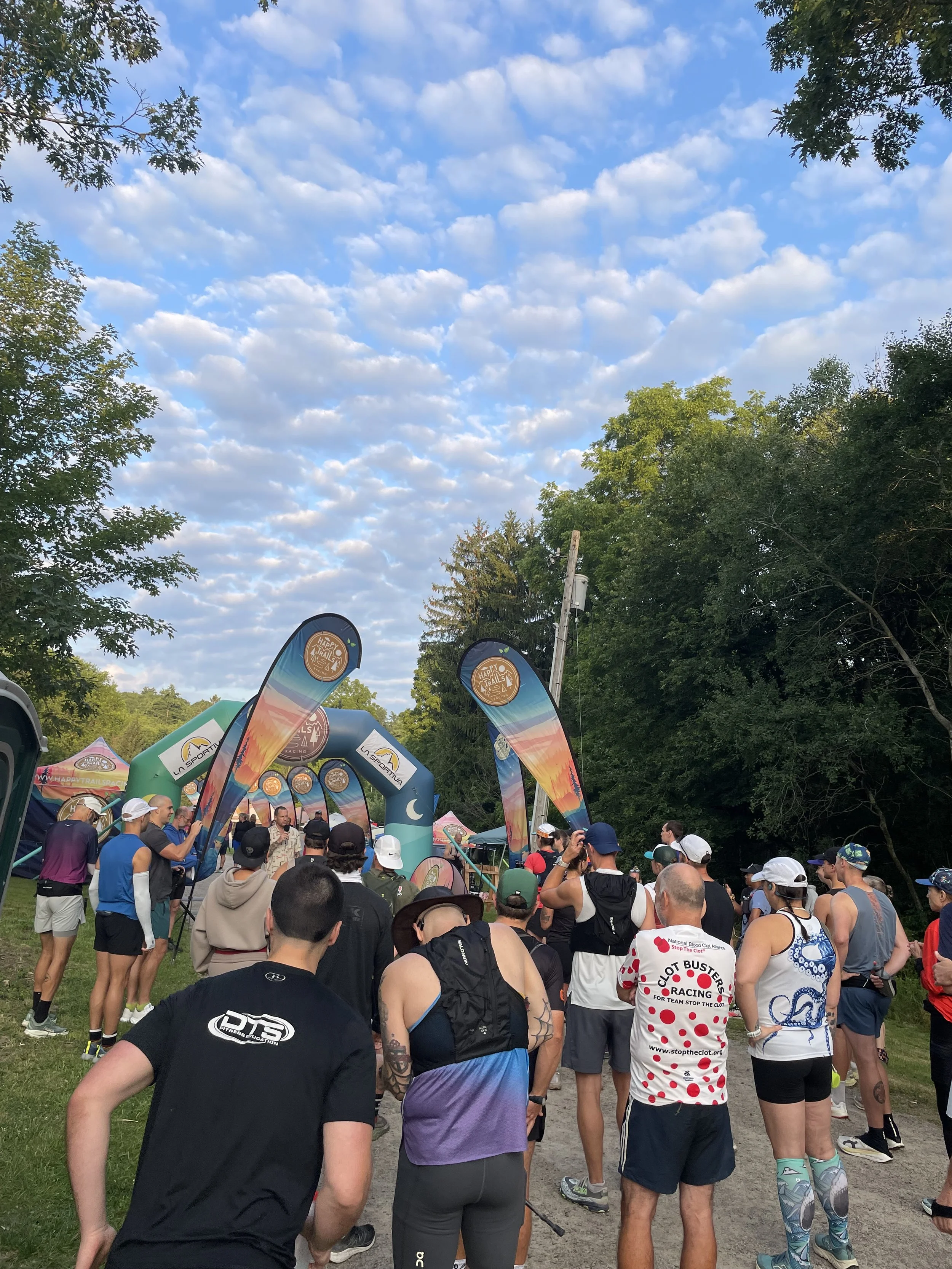 Crowd of runners gathered beneath colorful Happy Trails Racing banners at a wooded outdoor race event.