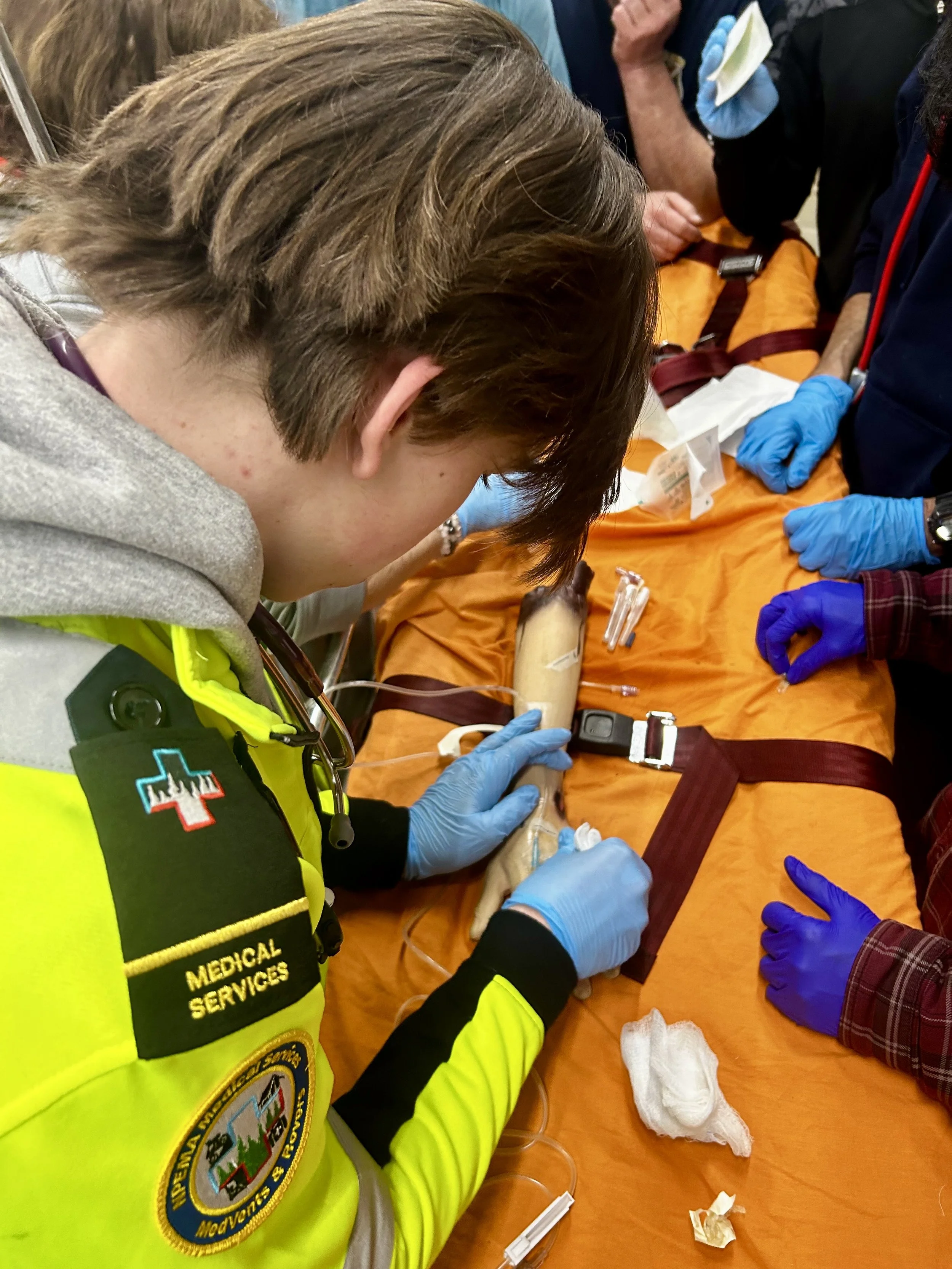 Paramedics in blue gloves treat an injured person on an orange stretcher during an emergency response.