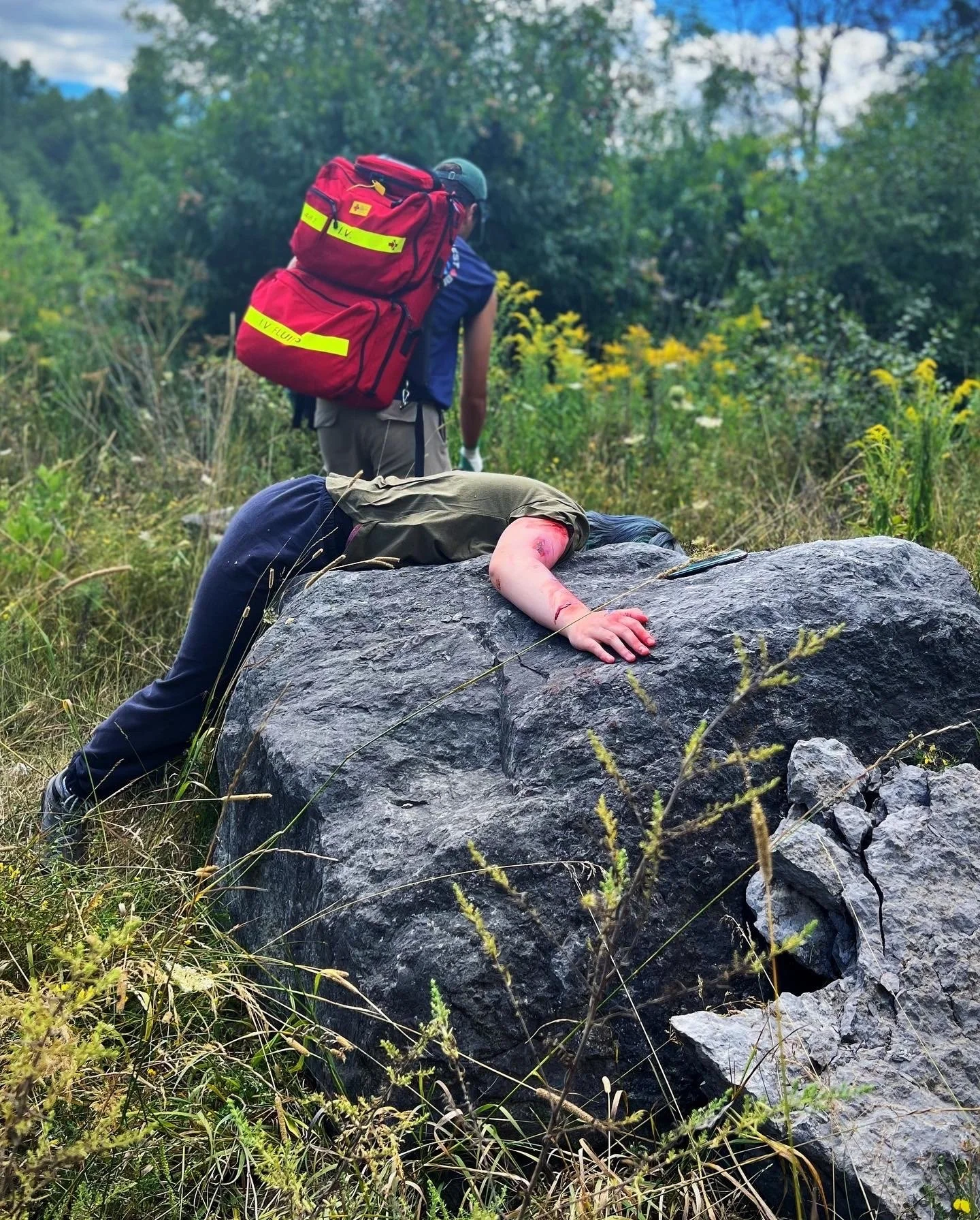 Search-and-rescue worker with a red medical backpack assists an injured hiker lying across a large rock outdoors.