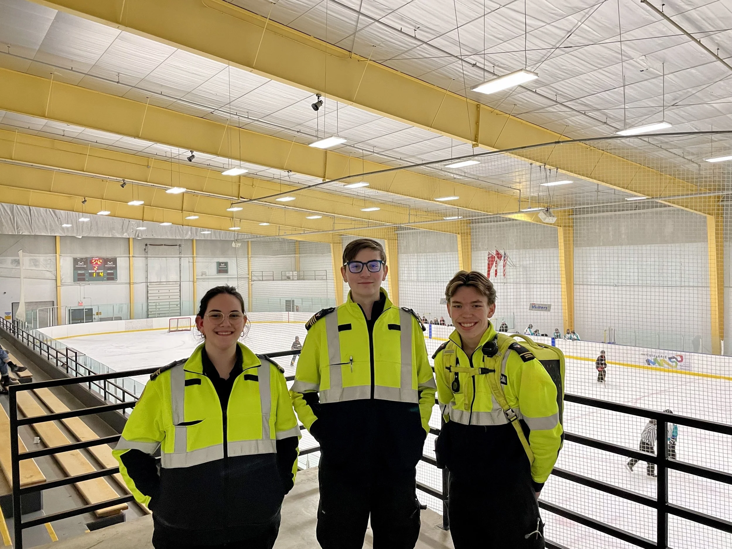 Three event staff members in high-visibility uniforms pose in front of an indoor ice hockey rink.