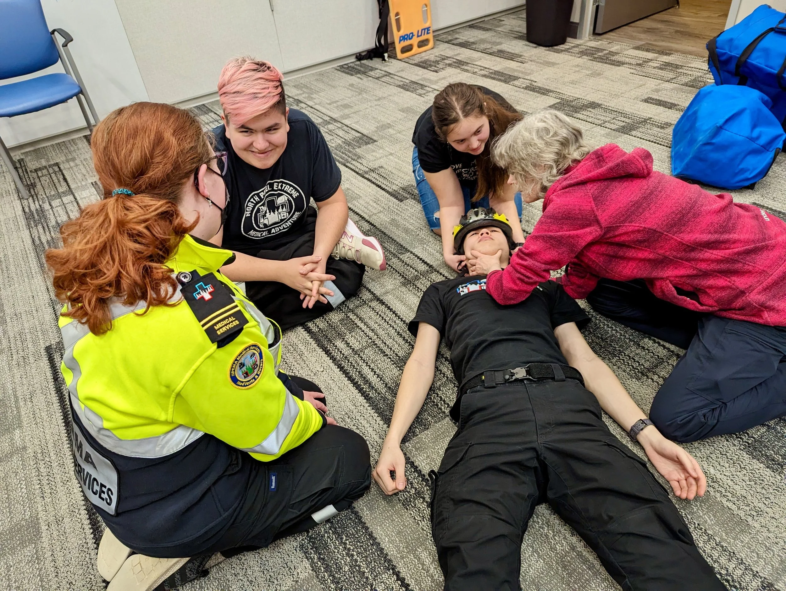 Emergency medical responders practice treating a person wearing a helmet on an office floor.