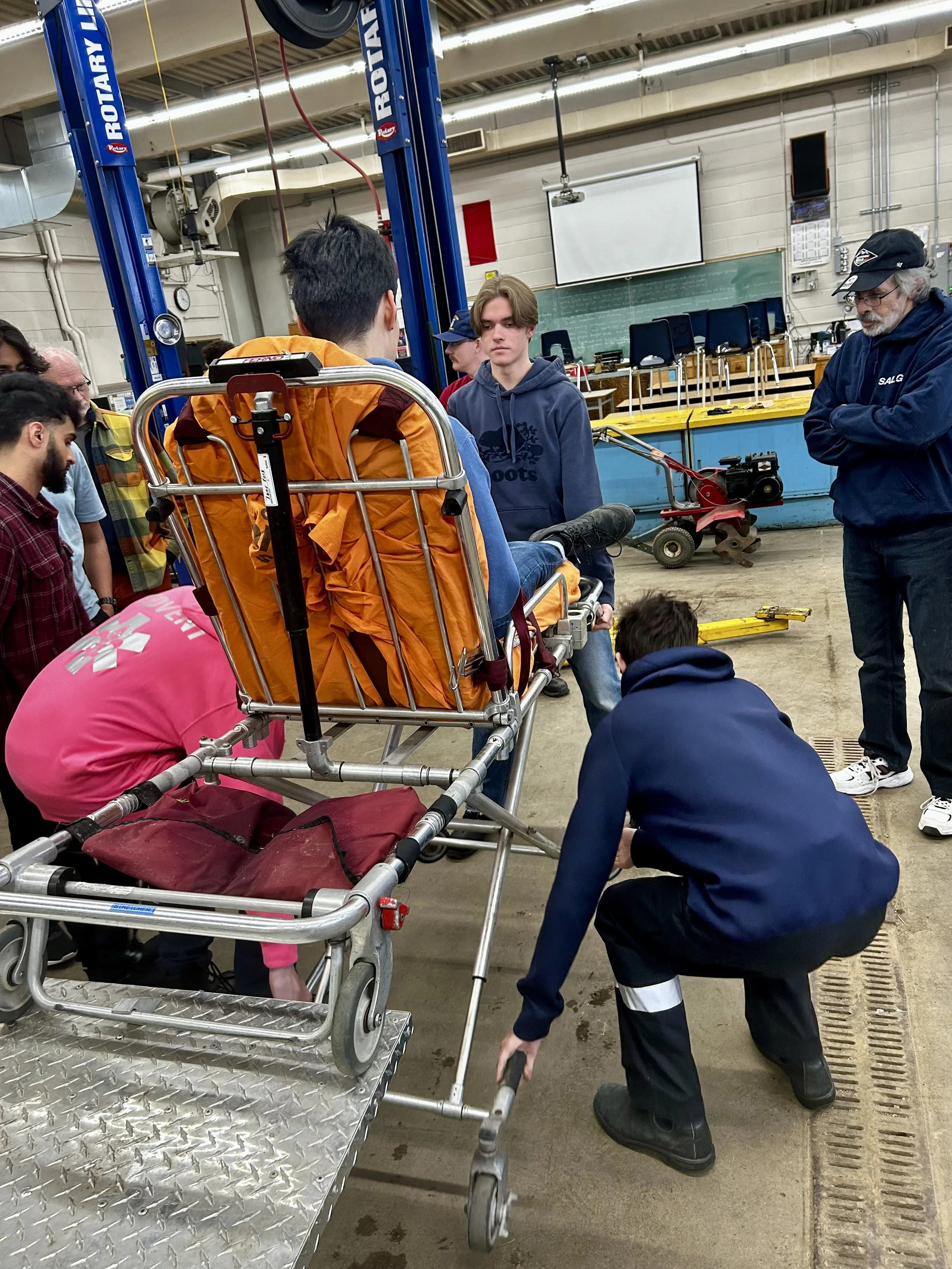 Group of people gathered around a stretcher in an automotive workshop with a Rotary vehicle lift.