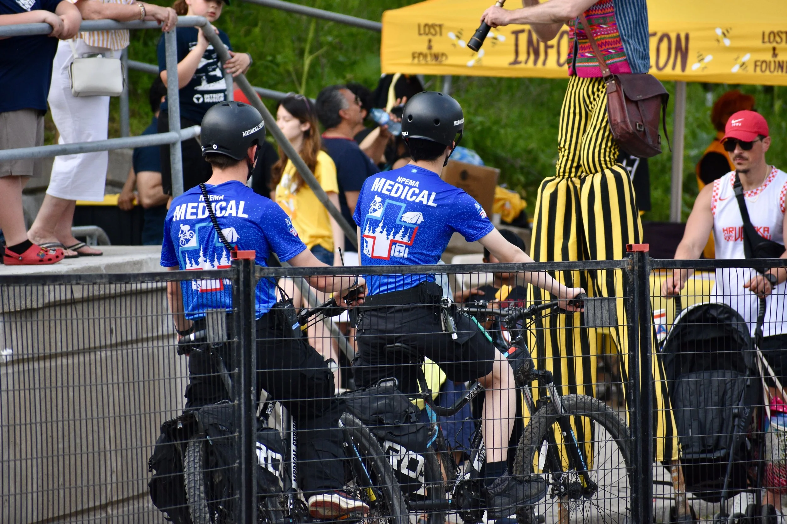 Medical volunteers on bicycles assist spectators at an outdoor event beside a person in a yellow-and-black striped costume.