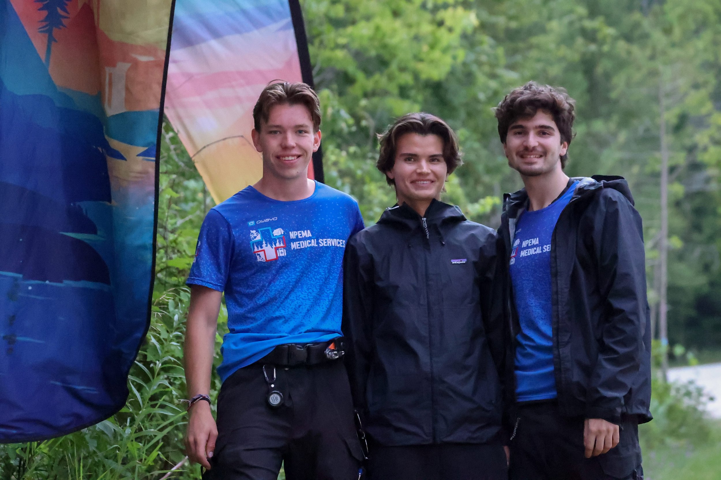 Three smiling npema medical responders pose outdoors beside a colorful mural; two wear blue NPEMA Medical Services shirts and one wears a black rain jacket.