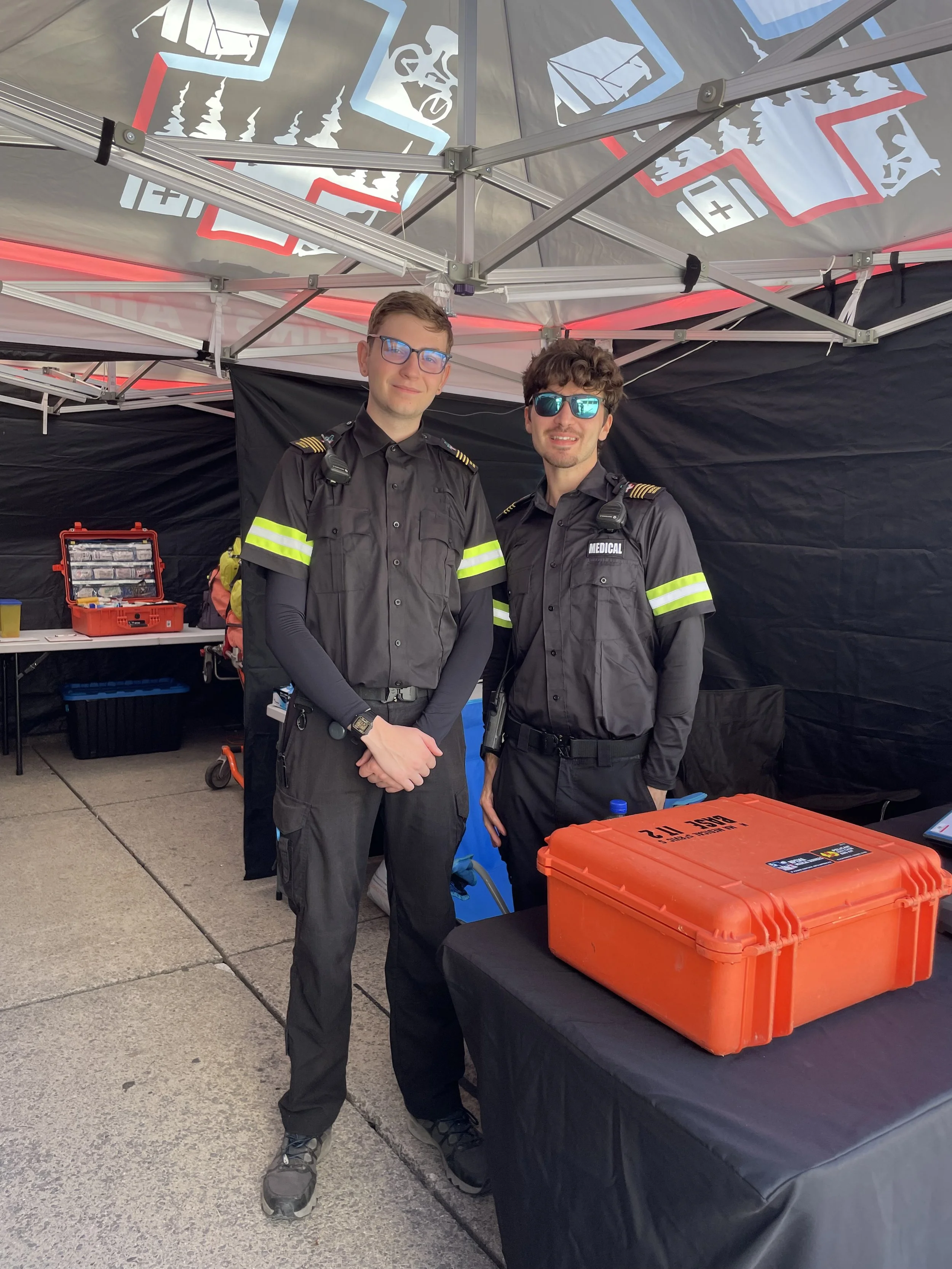 Two medical responders in uniform pose inside a tent beside orange emergency medical equipment cases.
