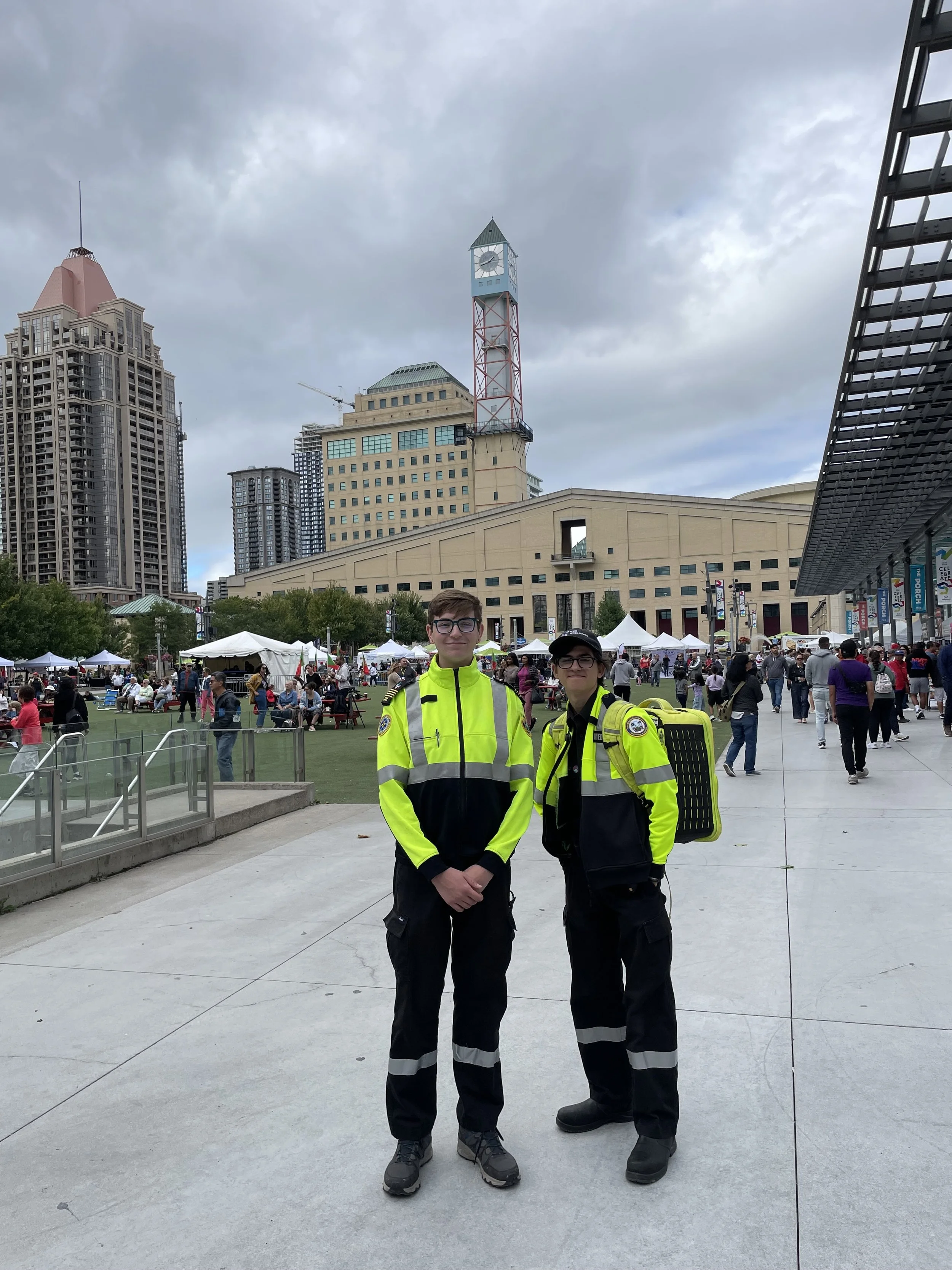 Two emergency medical responders in high-visibility uniforms at a busy outdoor festival, with city buildings and a clock tower in the background.