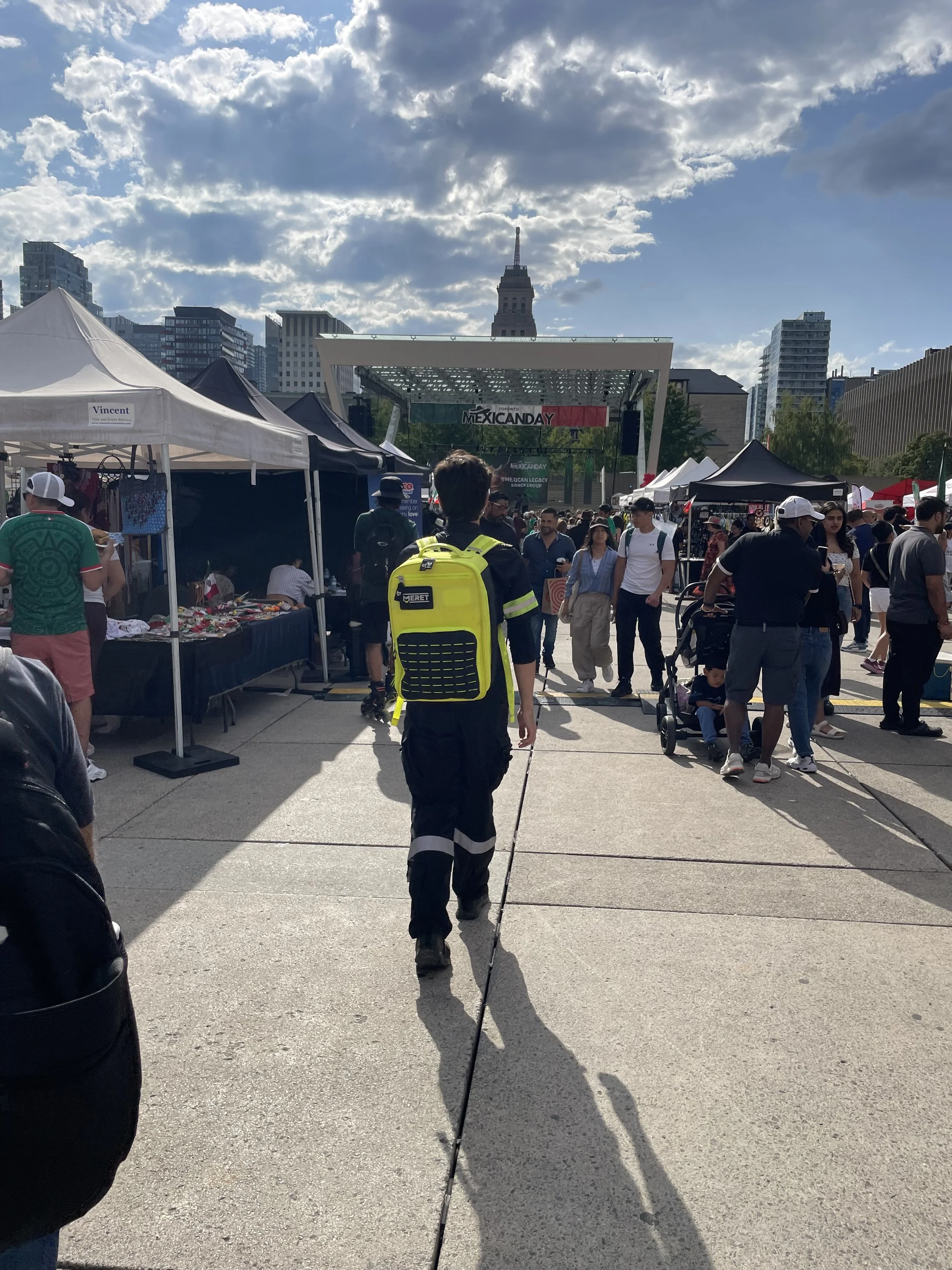 Crowded outdoor Mexican Day festival in Toronto with vendor tents, a stage, and downtown buildings under dramatic clouds