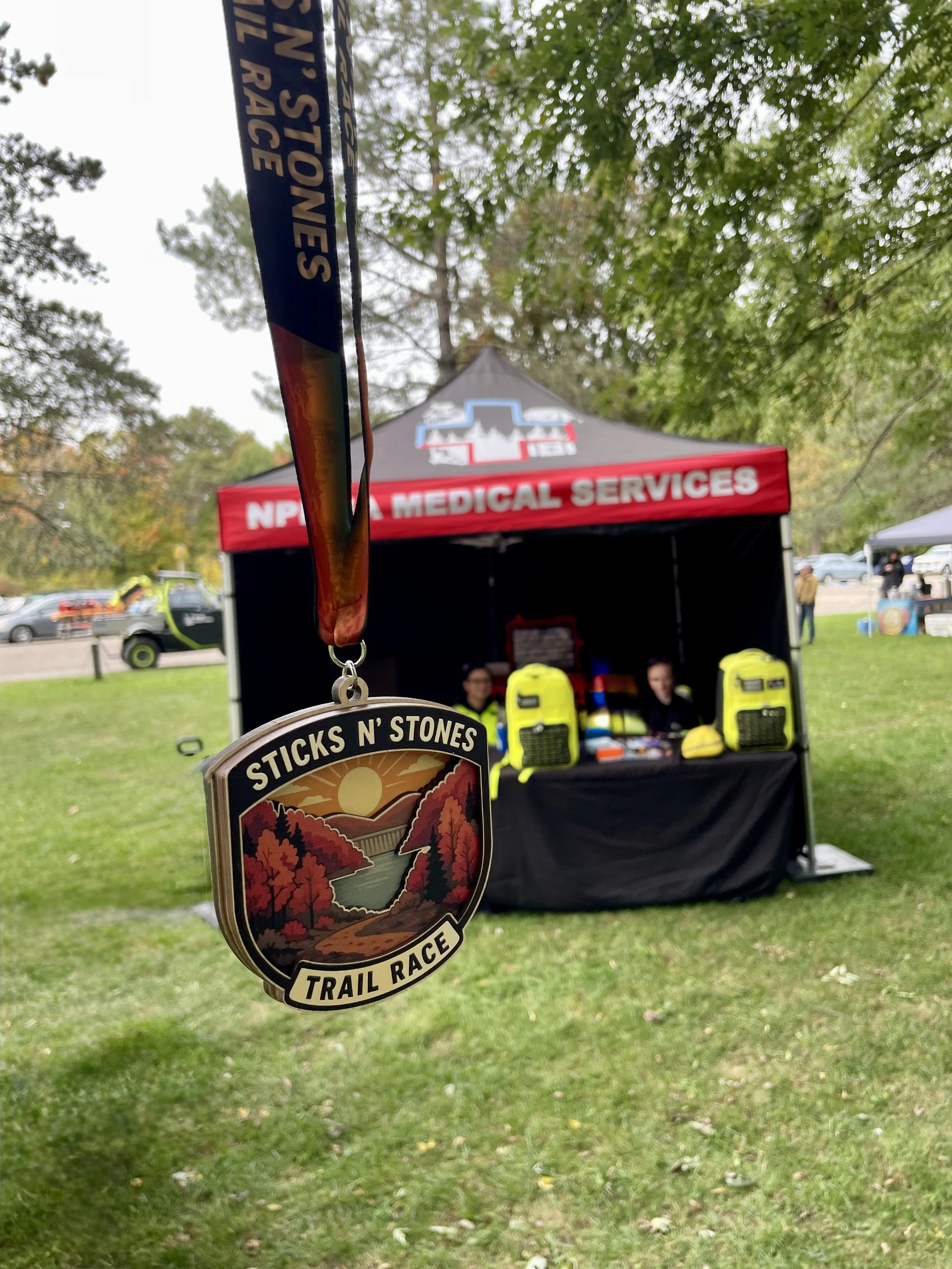 “Sticks N’ Stones Trail Race” medal hanging in front of an NPEMA medical services tent at an outdoor trail event.