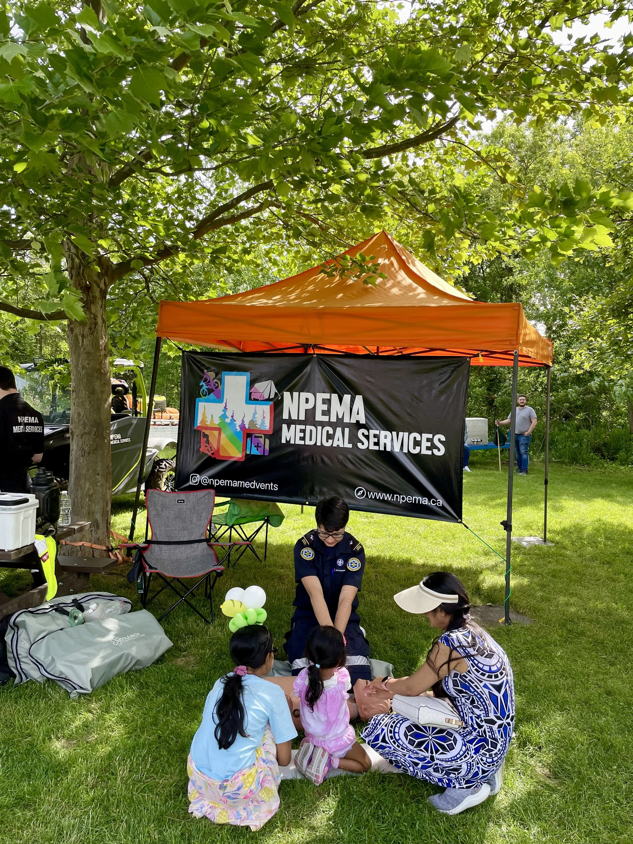 NPEMA Medical Services orange canopy at an outdoor community event, with a first responder, children, and families gathered under leafy trees.