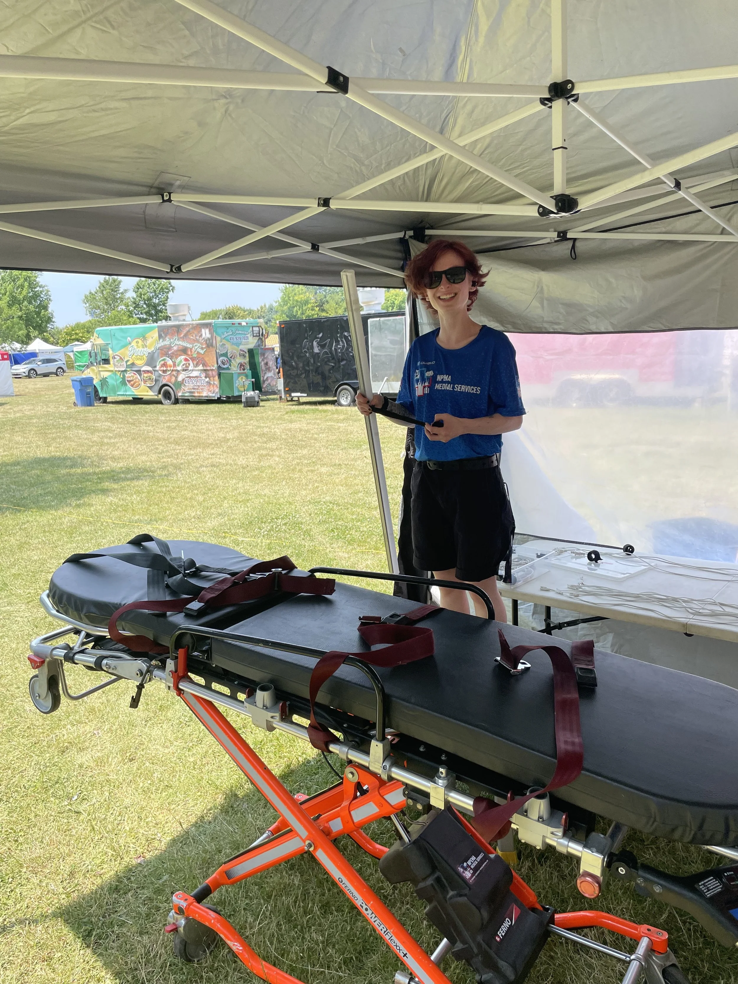 Medical volunteer in sunglasses standing beside a secured stretcher inside an outdoor event tent, with food trucks in the background.