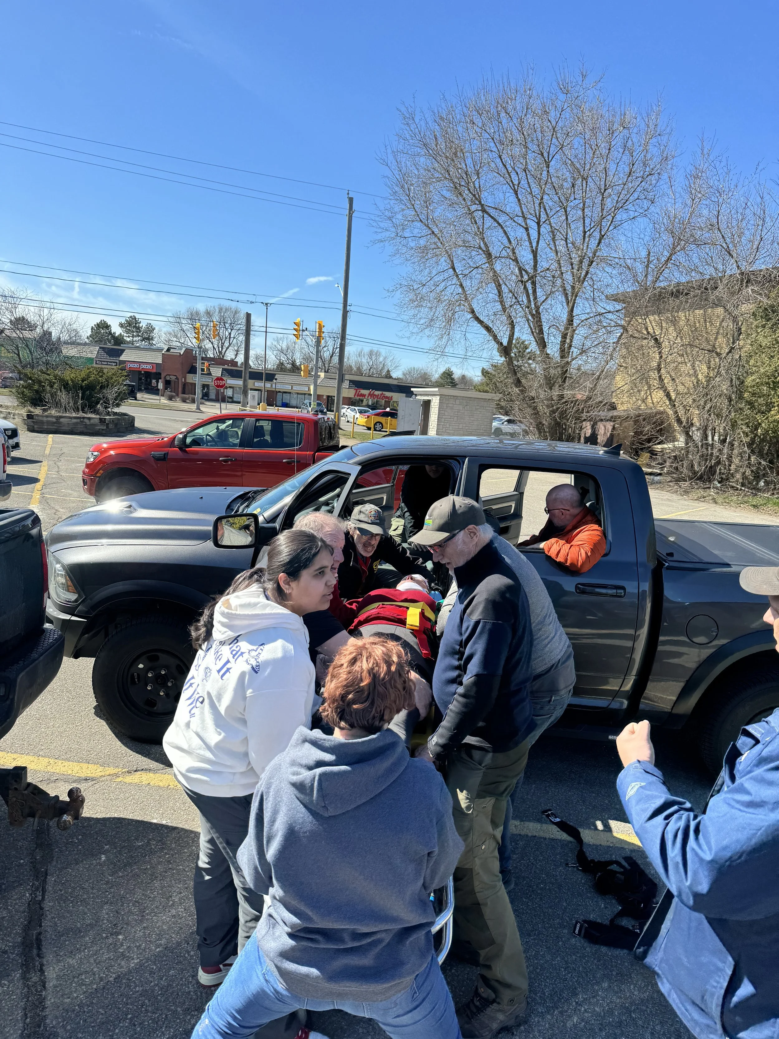 Group of people assisting an injured person beside parked vehicles in a shopping plaza parking lot, with Tim Hortons and traffic lights in the background.