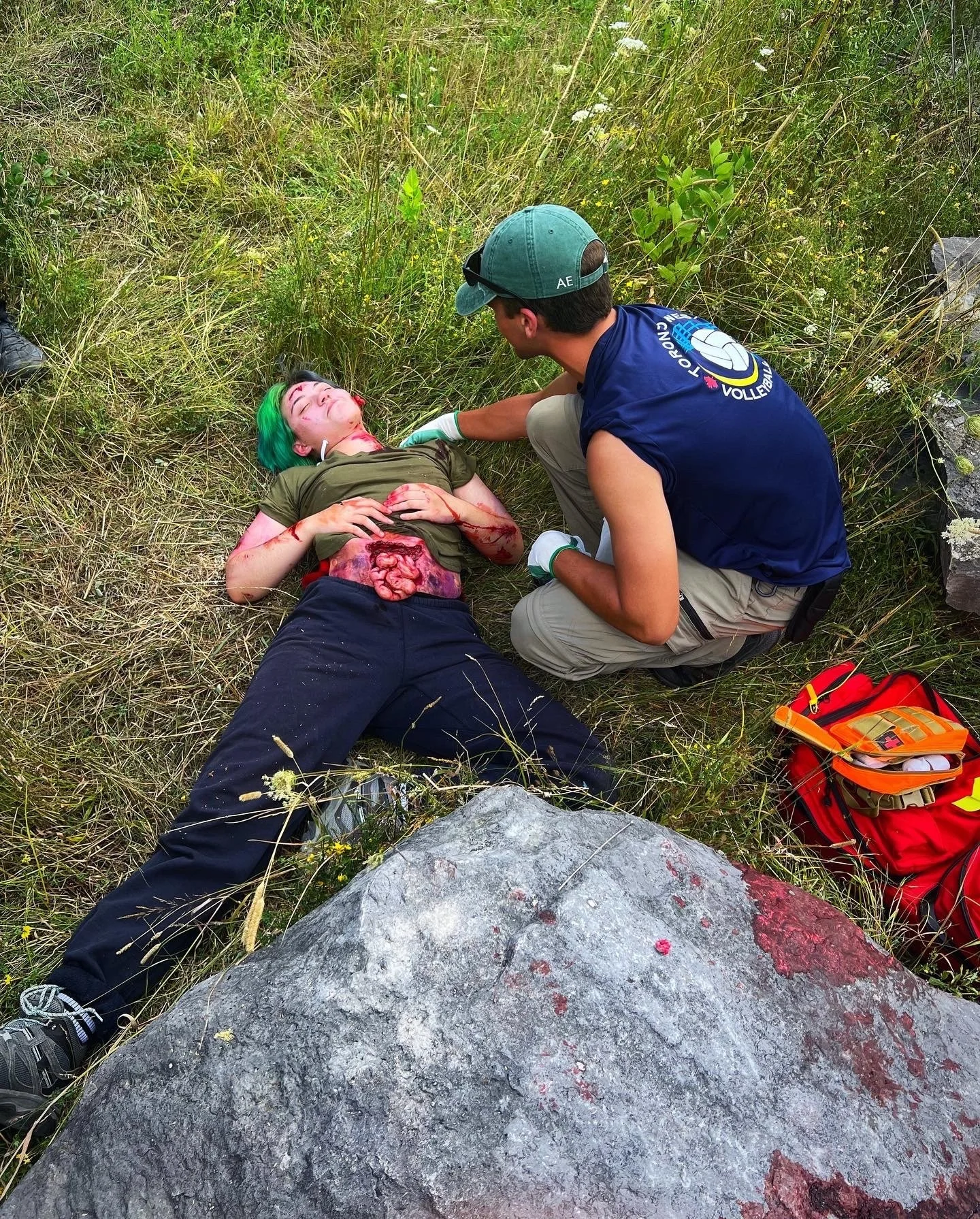 First responder treating an injured person lying beside a large rock in a grassy outdoor area, with a red emergency medical kit nearby