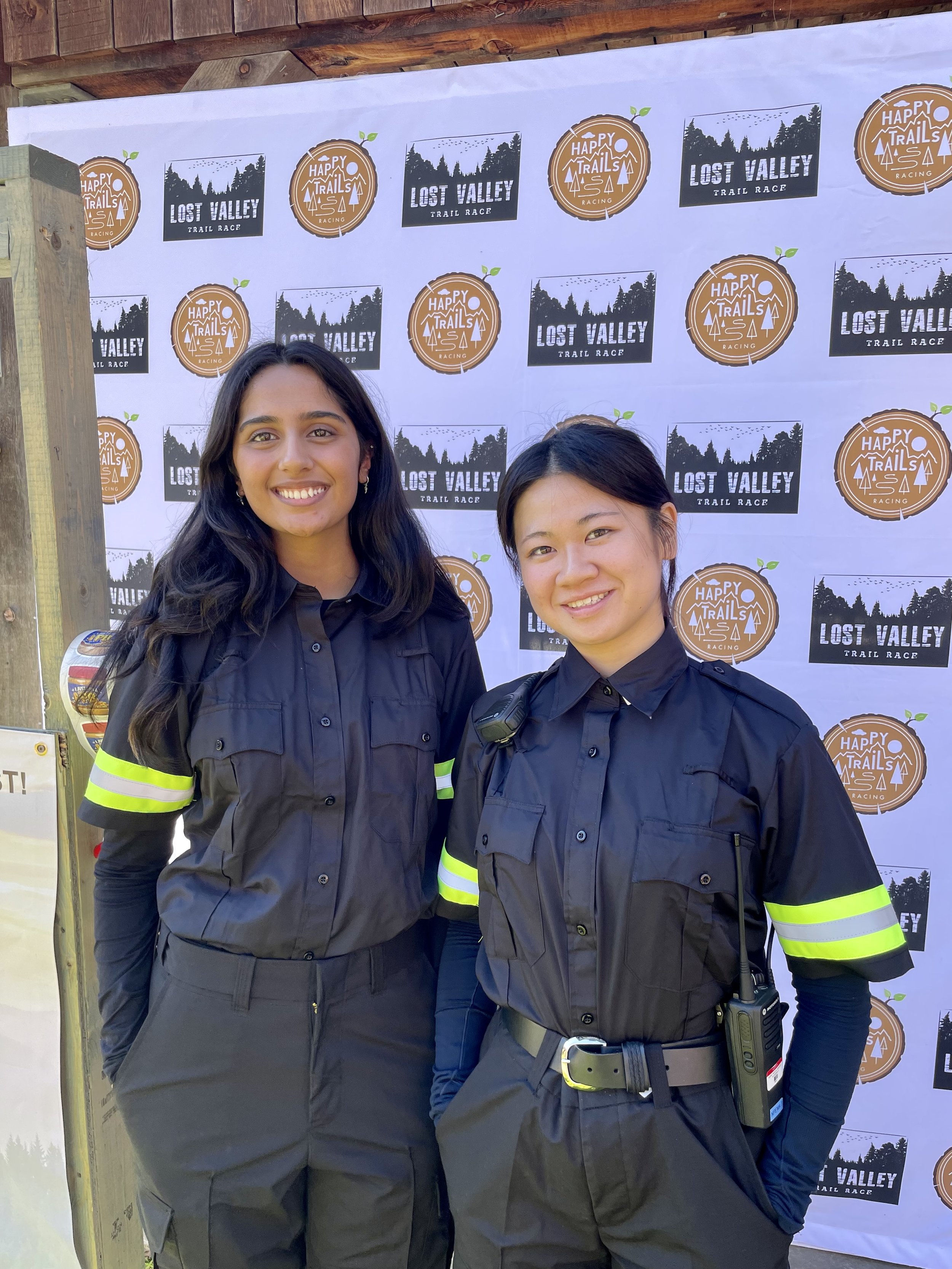 Two smiling npema medical responder staff members in black uniforms pose in front of a Lost Valley Trail Race and Happy Trails Racing backdrop.