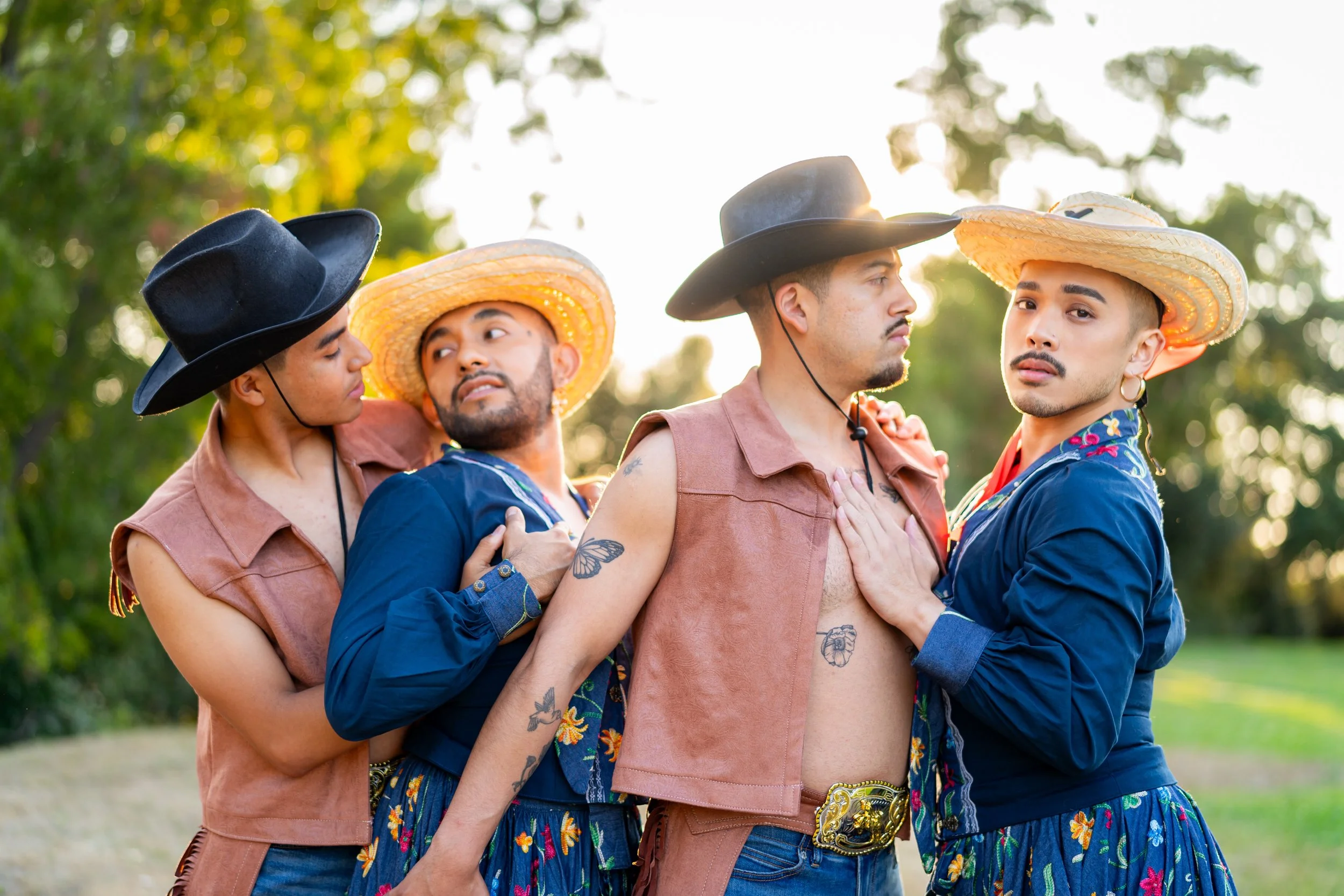 Four young men in cowboy hats and casual clothing, standing outdoors among trees, engaged in a close, affectionate moment, with one touching the chest of another while others look on.