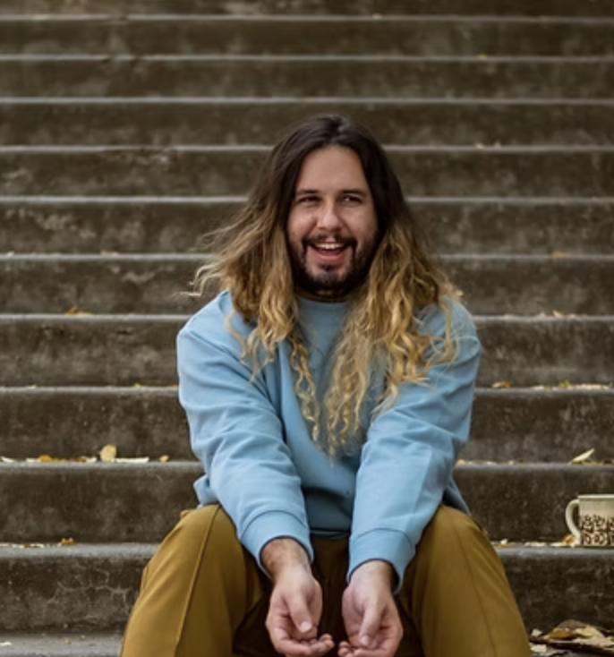 A man with long, wavy hair and a beard, smiling, sitting on outdoor concrete stairs, wearing a light blue sweatshirt and mustard-colored pants, with a mug on the stairs next to him.