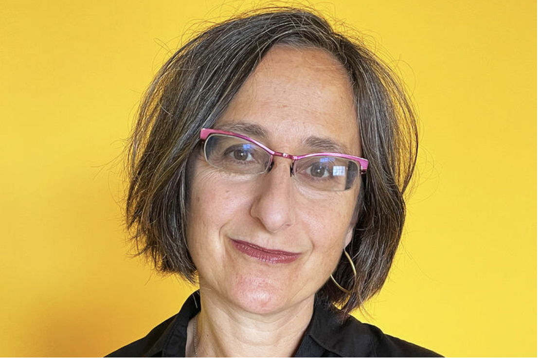 A woman with shoulder-length brown hair and pink glasses is smiling in front of a yellow background.
