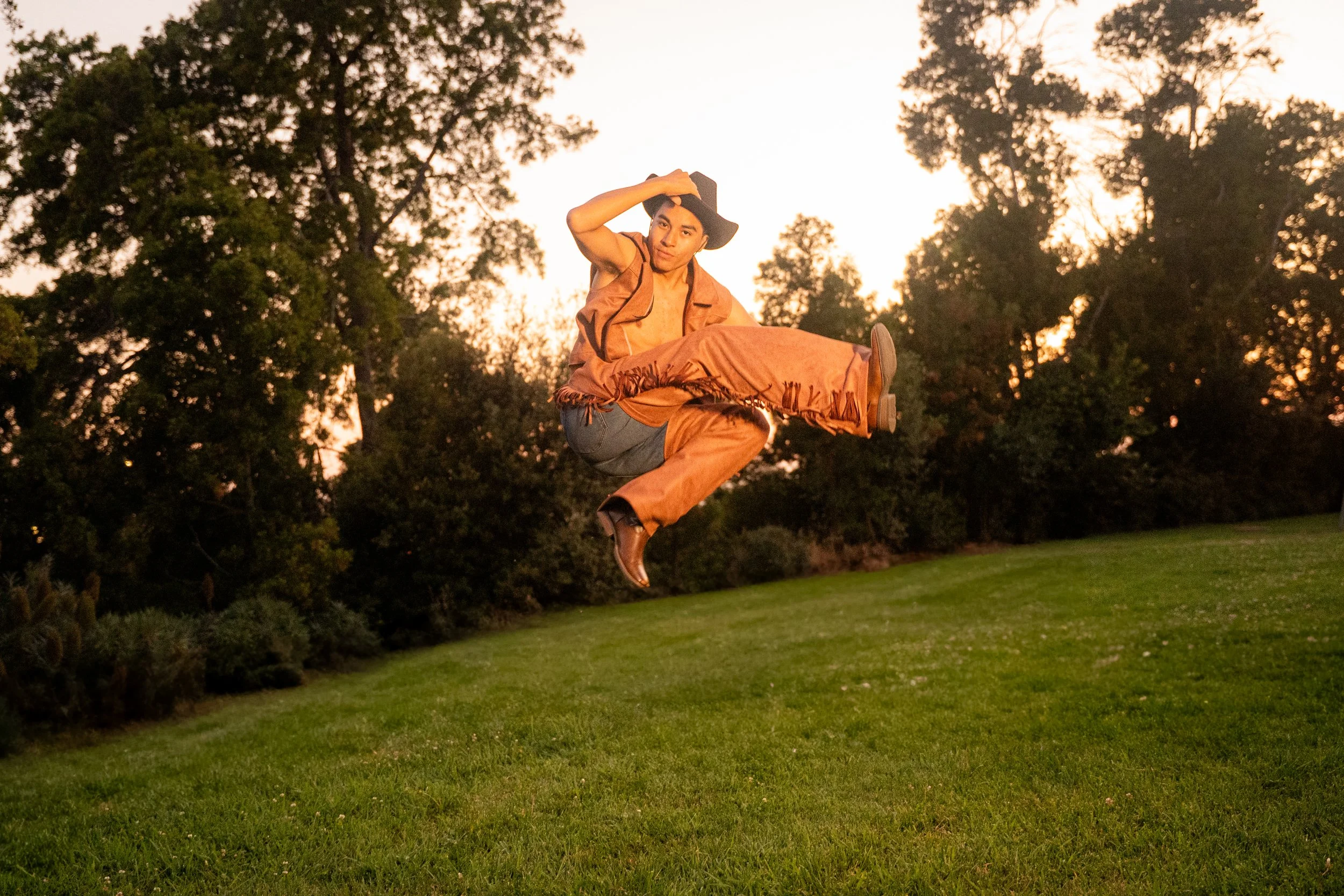 A young man dressed in cowboy attire, including a hat and fringed vest, jumps into the air outdoors during sunset, with trees and grass in the background.
