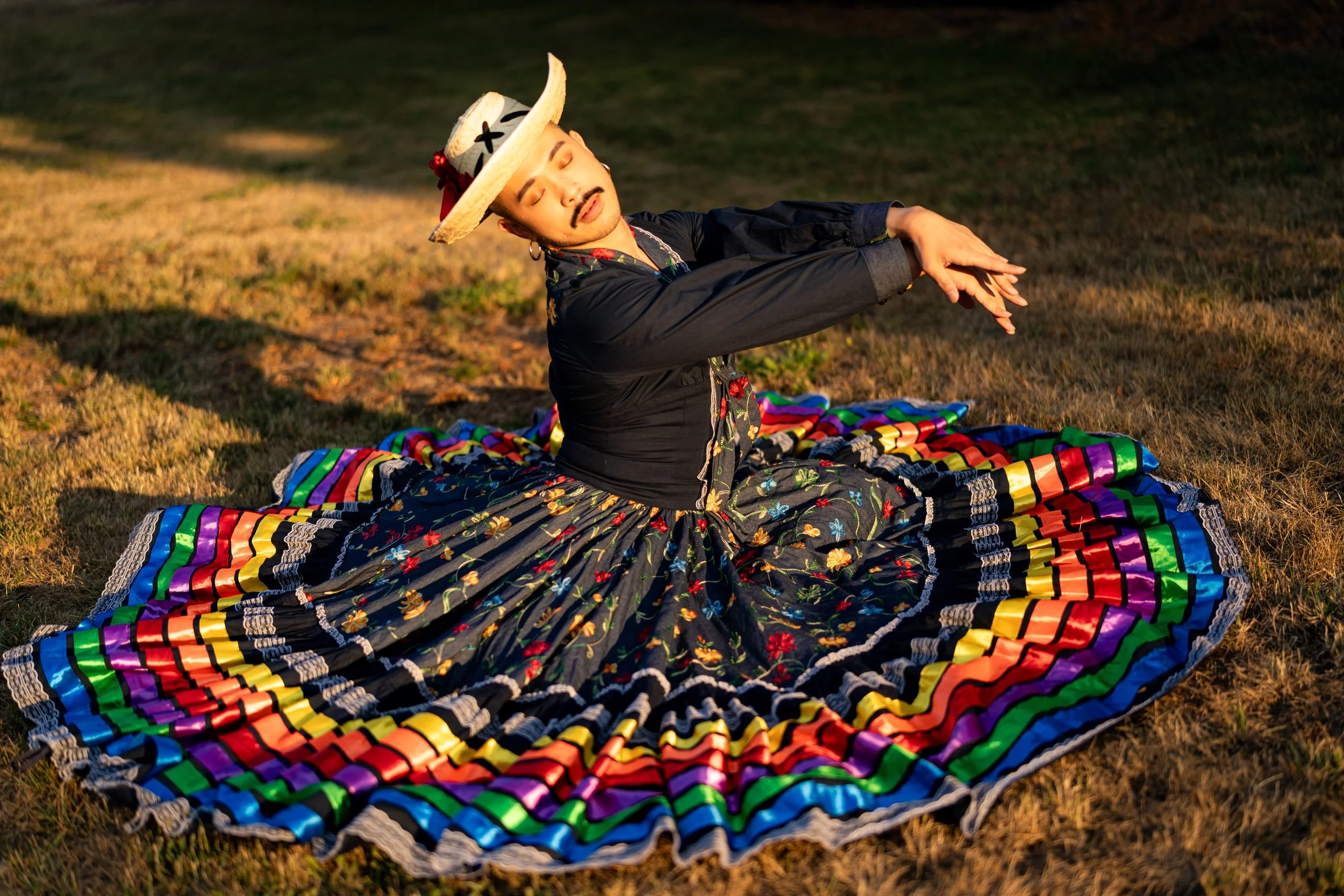 Person practicing yoga outdoors on a colorful, patterned blanket in the evening, wearing a black floral skirt, a black top, and a traditional hat, with eyes closed and arms extended forward.
