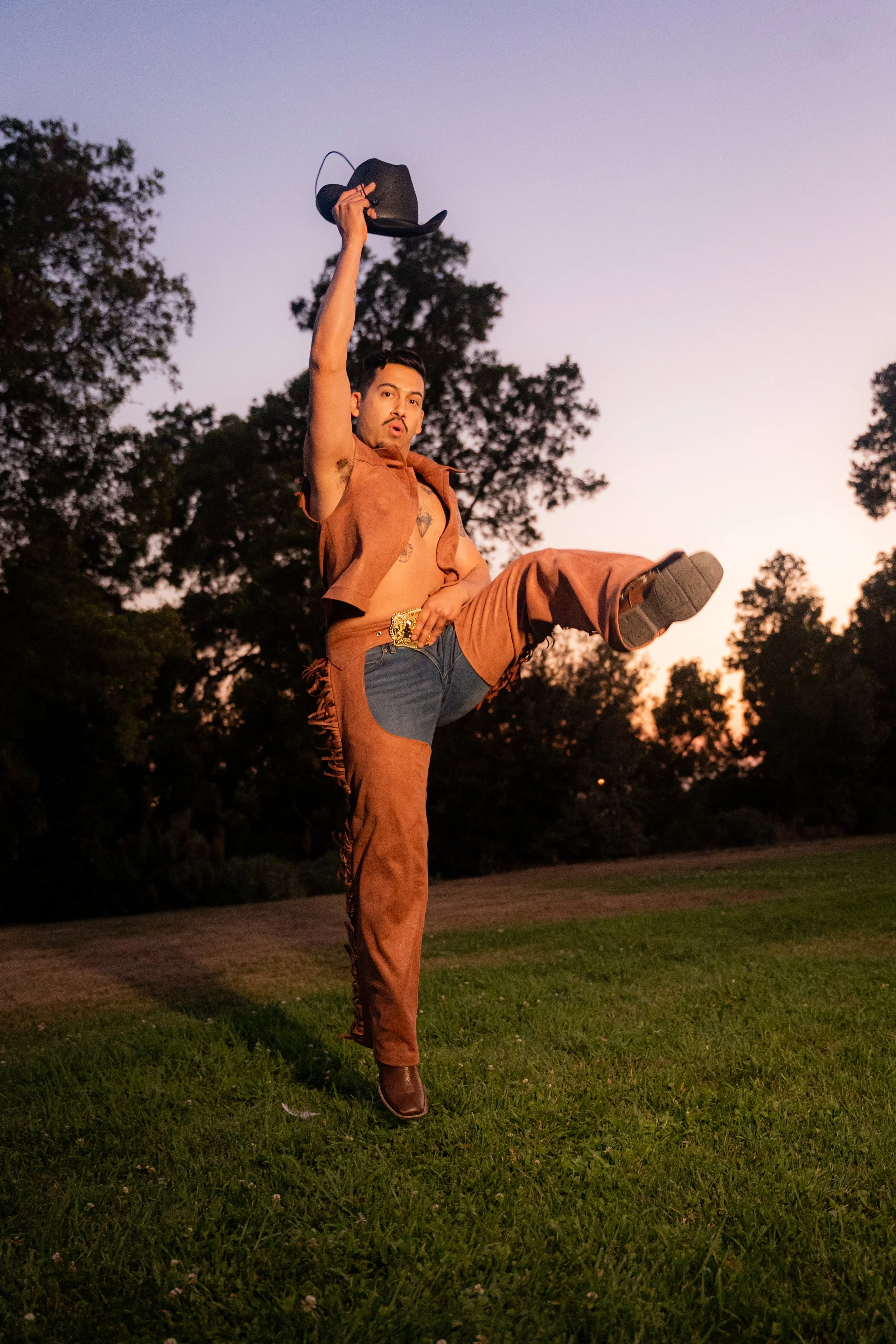 A man dressed as a cowboy balancing on one leg in a grassy field during sunset, wearing a brown vest and pants, holding a black cowboy hat in the air.