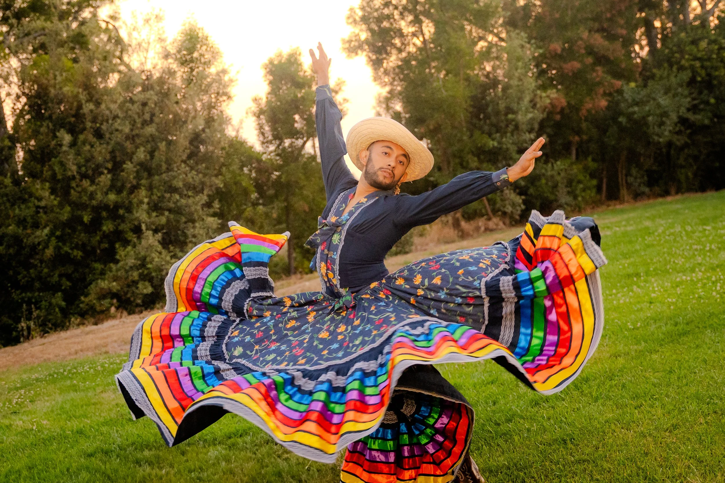 A man wearing a hat, dark shirt, and patterned skirt performs a dance outdoors during sunset, with trees in the background and colorful fabric flowing around him.
