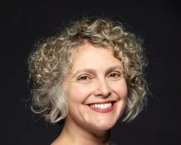 Close-up of a smiling woman with curly gray hair against a black background.
