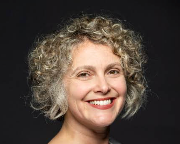Close-up portrait of a middle-aged woman with curly blonde hair smiling against a dark background.