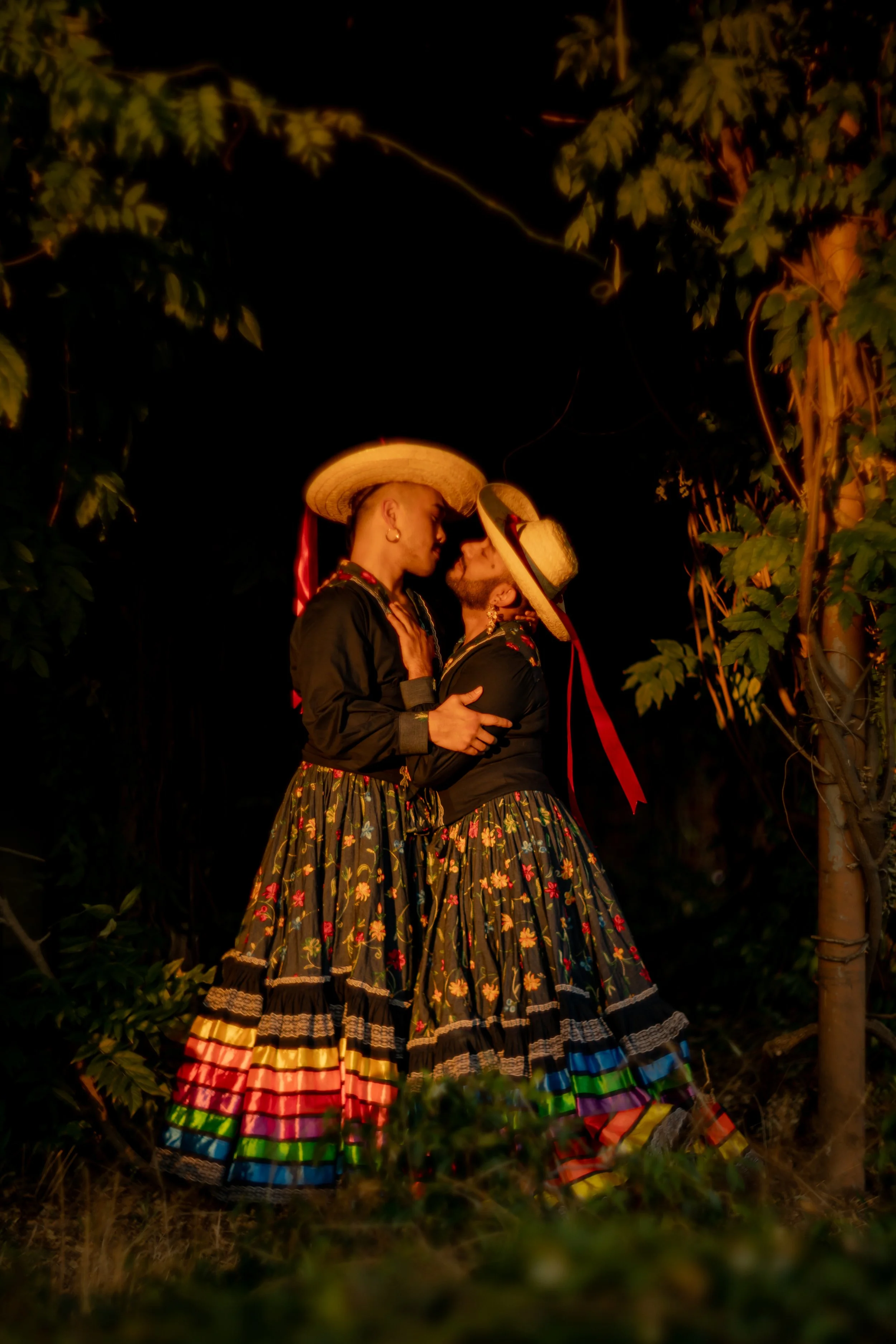 Two individuals dressed in traditional Mexican attire, wearing wide-brimmed sombreros, engaged in an intimate moment during nighttime outdoors, surrounded by trees with dark night sky background.
