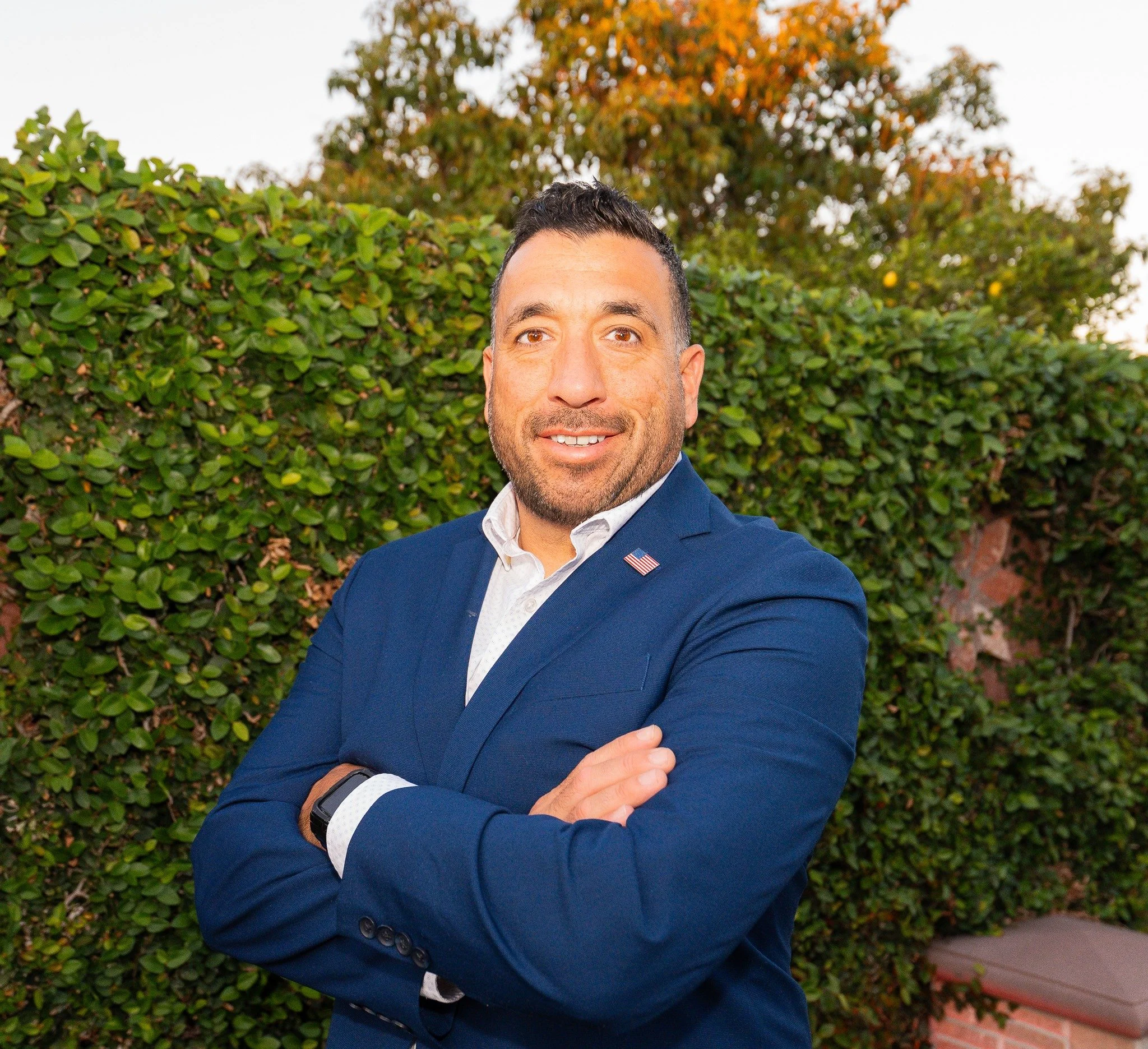 Carlos Yado in a navy blazer, standing outdoors with his arms crossed.