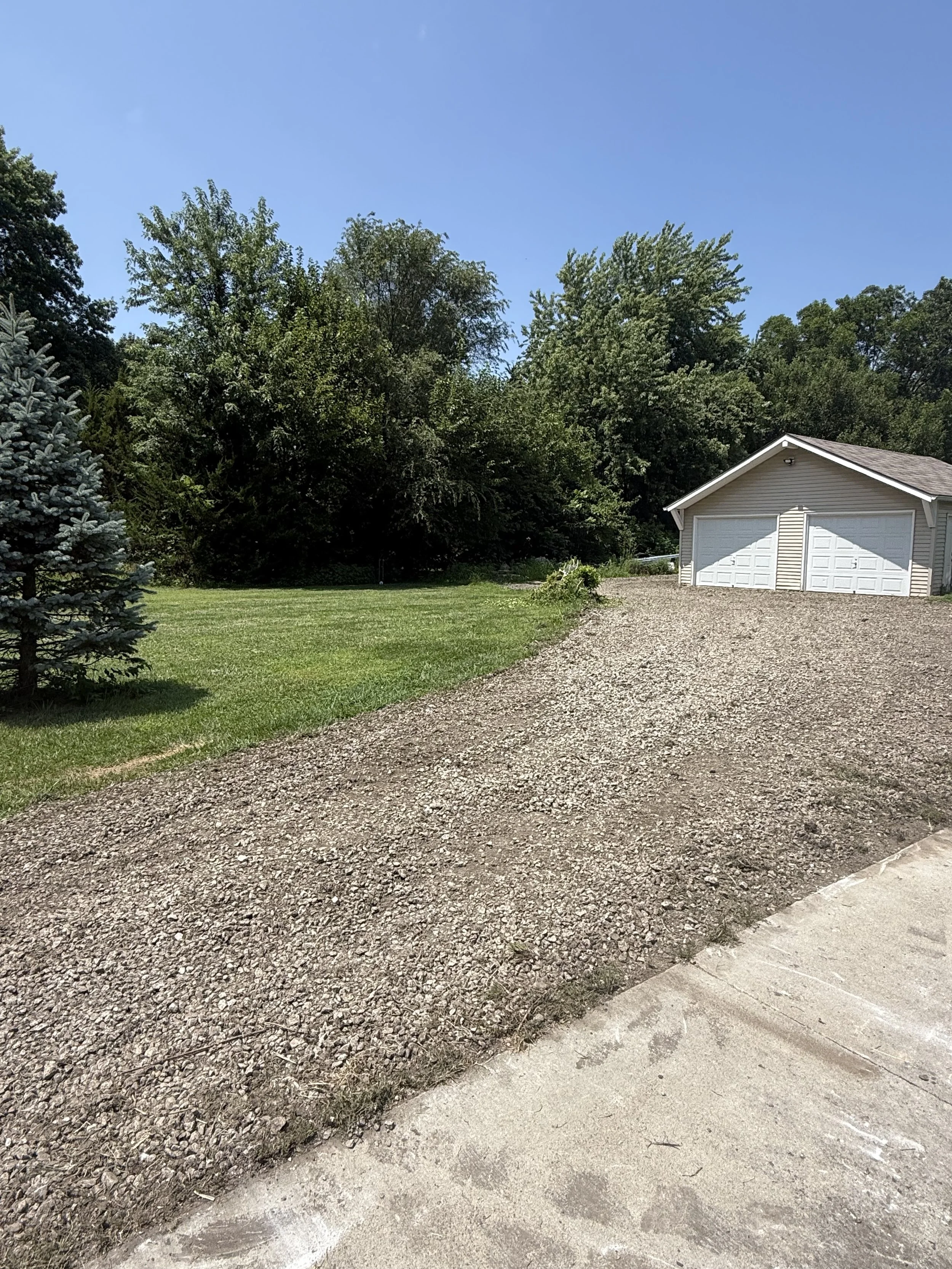 A landscape showing a gravel driveway and a grass lawn with a white garage, trees, and a clear blue sky.