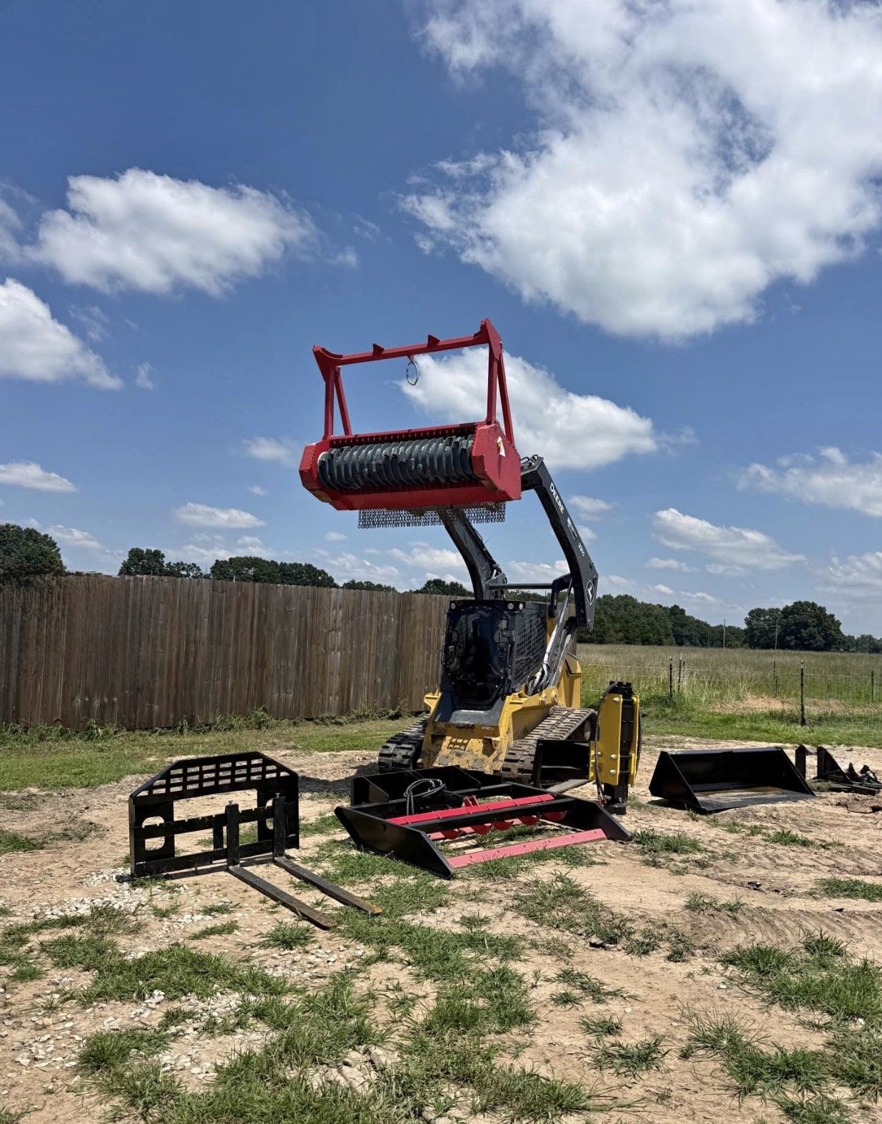 A mini excavator with a red attachment raised in the air, on a construction site with an open field and a wooden fence in the background, under a partly cloudy sky.